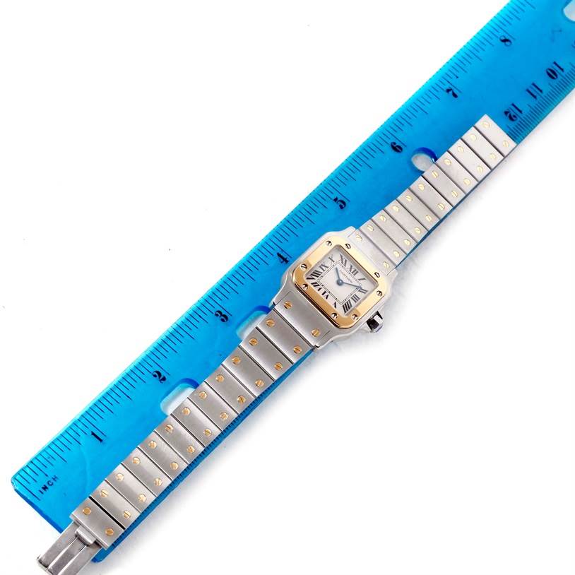 The Santos model by Cartier is shown from above, including the watch face, bracelet, and a blue ruler for scale.