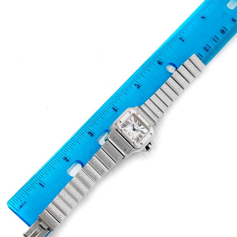 The image shows an overhead view of the Cartier Santos W20054D6 Women
s Stainless Steel Silver Dial W20054D6 Women
s Stainless Steel Silver Dial watch, including its face and full metal bracelet, alongside a blue ruler for scale.