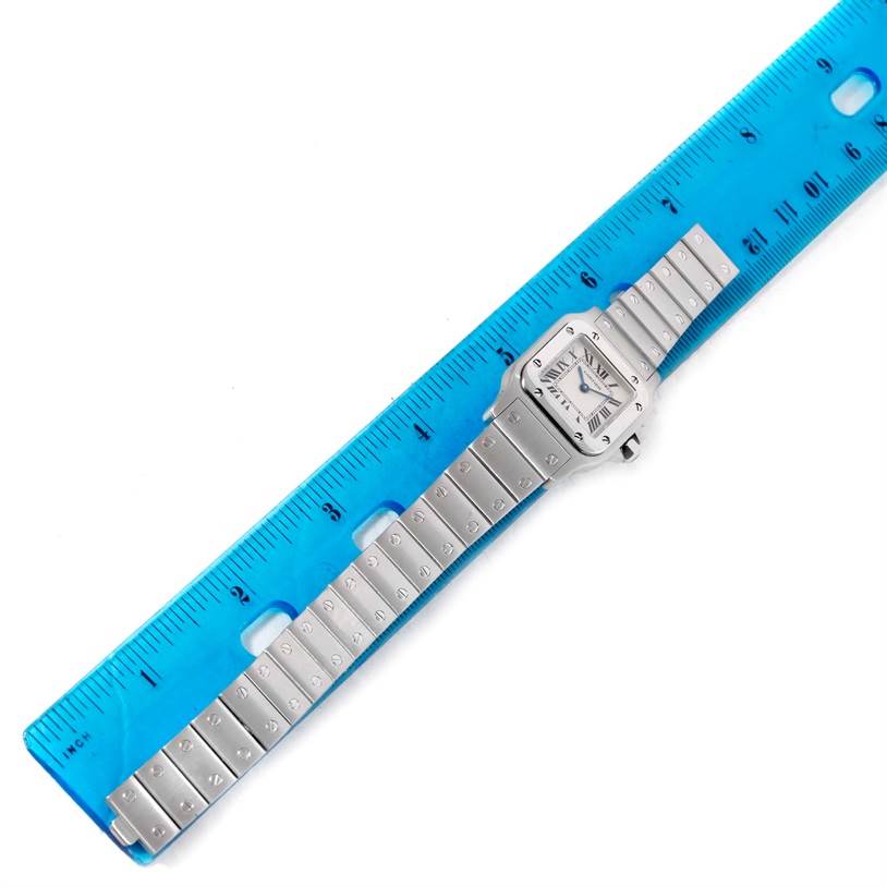 The image shows a top-down view of a Cartier Santos W20056D6 Women
s Stainless Steel Silver Dial W20056D6 Women
s Stainless Steel Silver Dial model watch on a blue ruler, displaying the watch face and full metal band.