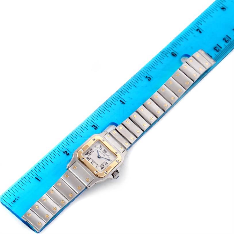 The image shows a top-down view of a Cartier Santos W20012C4 Women
s Stainless Steel Silver Dial W20012C4 Women
s Stainless Steel Silver Dial watch with its stainless steel and gold bracelet next to a ruler.