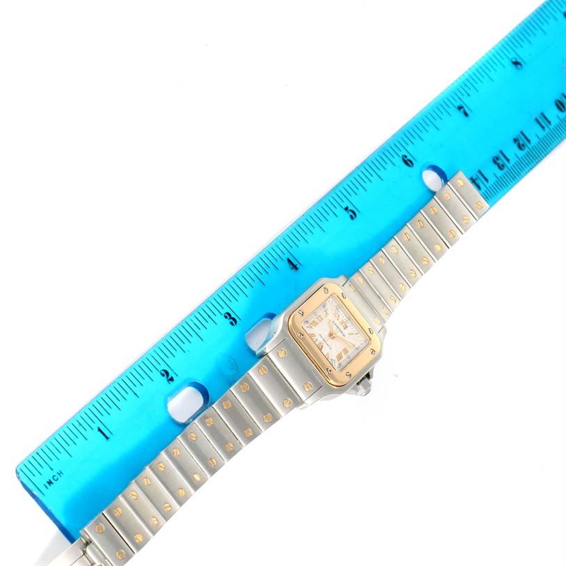 The Santos model by Cartier is shown from a top-down angle, alongside a blue ruler, highlighting the watch face and bracelet.