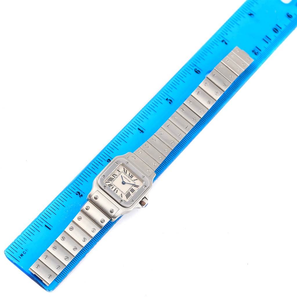 The Cartier Santos W20056D6 Women
s Stainless Steel Silver Dial W20056D6 Women
s Stainless Steel Silver Dial watch is shown from a top-down angle, including the watch face and metal strap, laid next to a blue ruler.