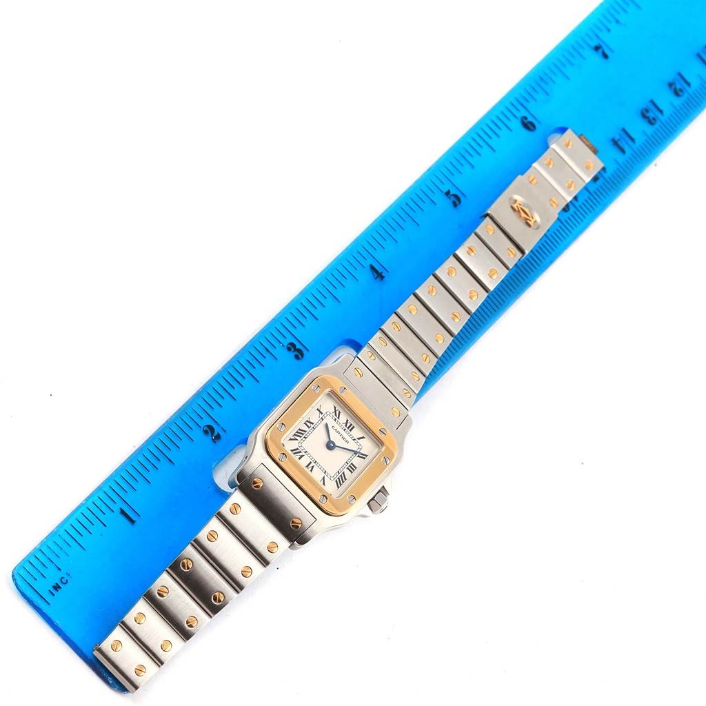 The Santos model by Cartier is shown lying flat with a blue ruler beside it, displaying the bracelet and watch face.