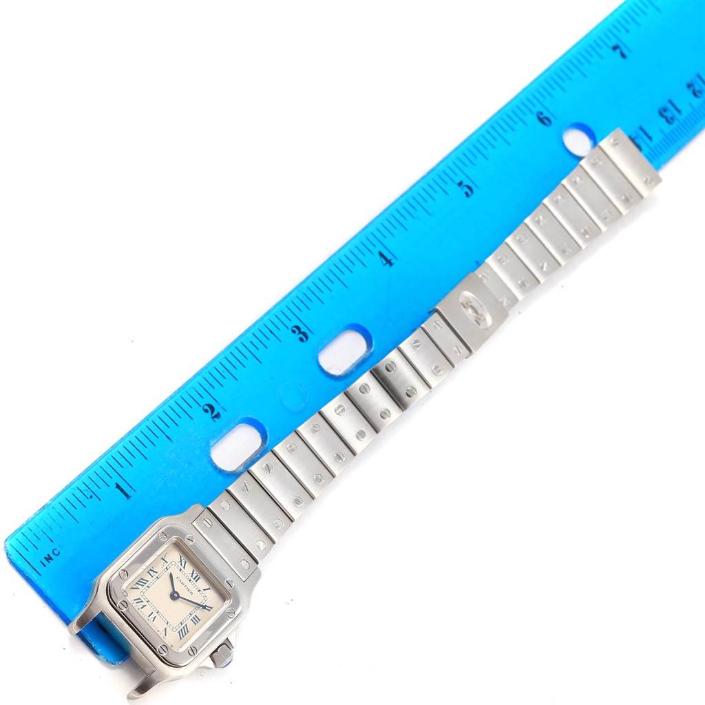 This image shows a Cartier Santos 07791 Women's Stainless Steel Silver Dial 07791 Women's Stainless Steel Silver Dial watch laid flat next to a blue ruler, displaying its face and metal bracelet.