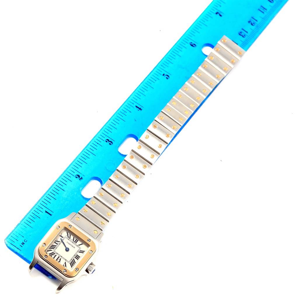 The image shows a top-down view of a Cartier Santos W20012C4 Women's Stainless Steel Silver Dial W20012C4 Women's Stainless Steel Silver Dial watch with its face, bezel, and bracelet placed next to a blue ruler.