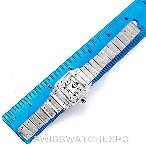 The image shows an overhead view of a Cartier Santos W20056D6 Women
s Stainless Steel Silver Dial W20056D6 Women
s Stainless Steel Silver Dial watch, with a stainless steel band and white dial, placed next to a blue ruler for scale.