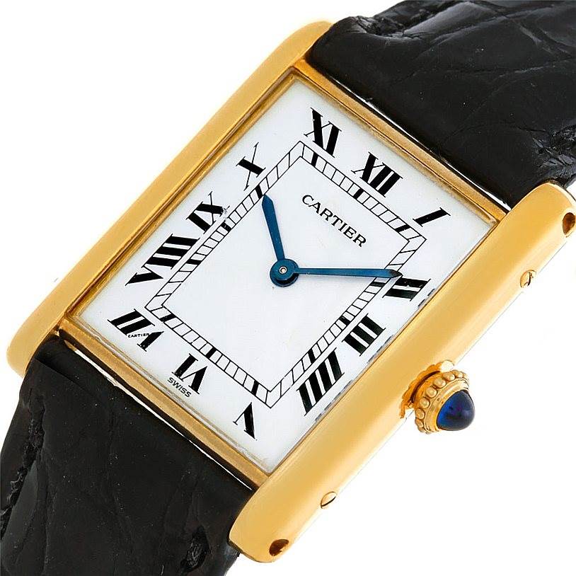 The image shows a Cartier Tank Classic Men's Yellow Gold White Dial Men's Yellow Gold White Dial watch angled to display its face, gold casing, black strap, and blue cabochon crown.