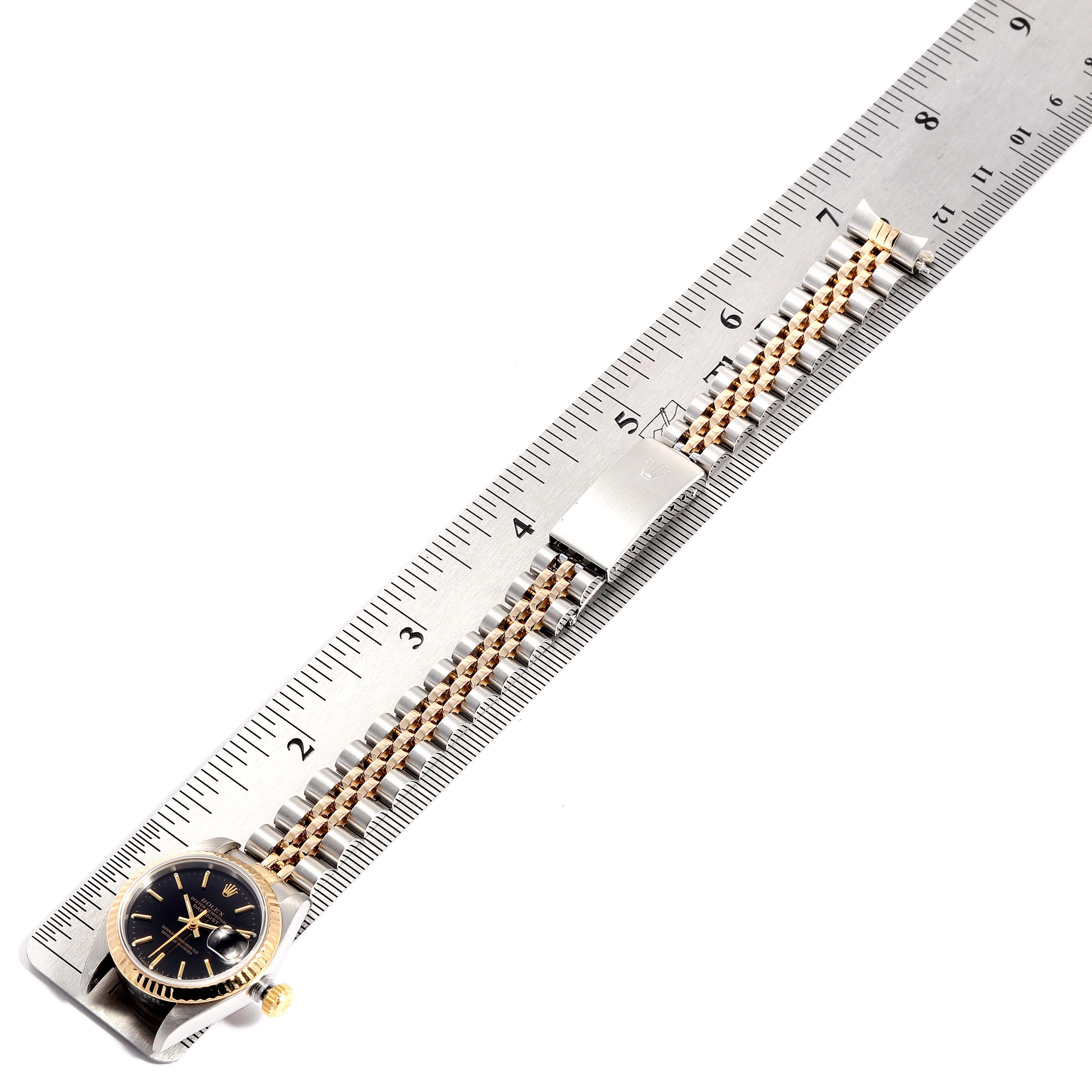 The image shows a Rolex Datejust 69173 Women
s Steel and Gold (two tone) Black Dial 69173 Women
s Steel and Gold (two tone) Black Dial watch laid flat beside a ruler, highlighting its bracelet and clasp.