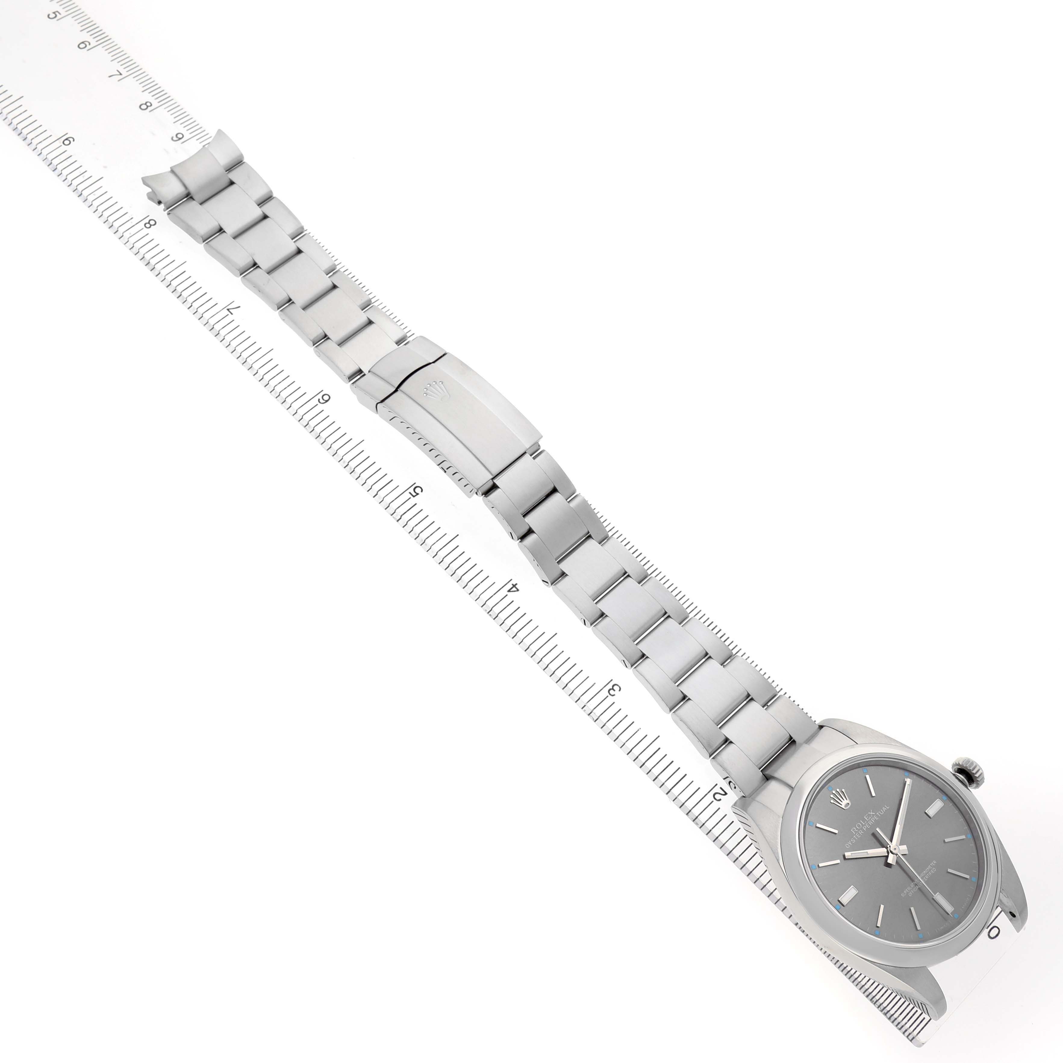 The image shows a Rolex Oyster Perpetual 114300 Men's Stainless Steel Grey Dial watch diagonally, displaying the watch face, bracelet, and clasp alongside a ruler.