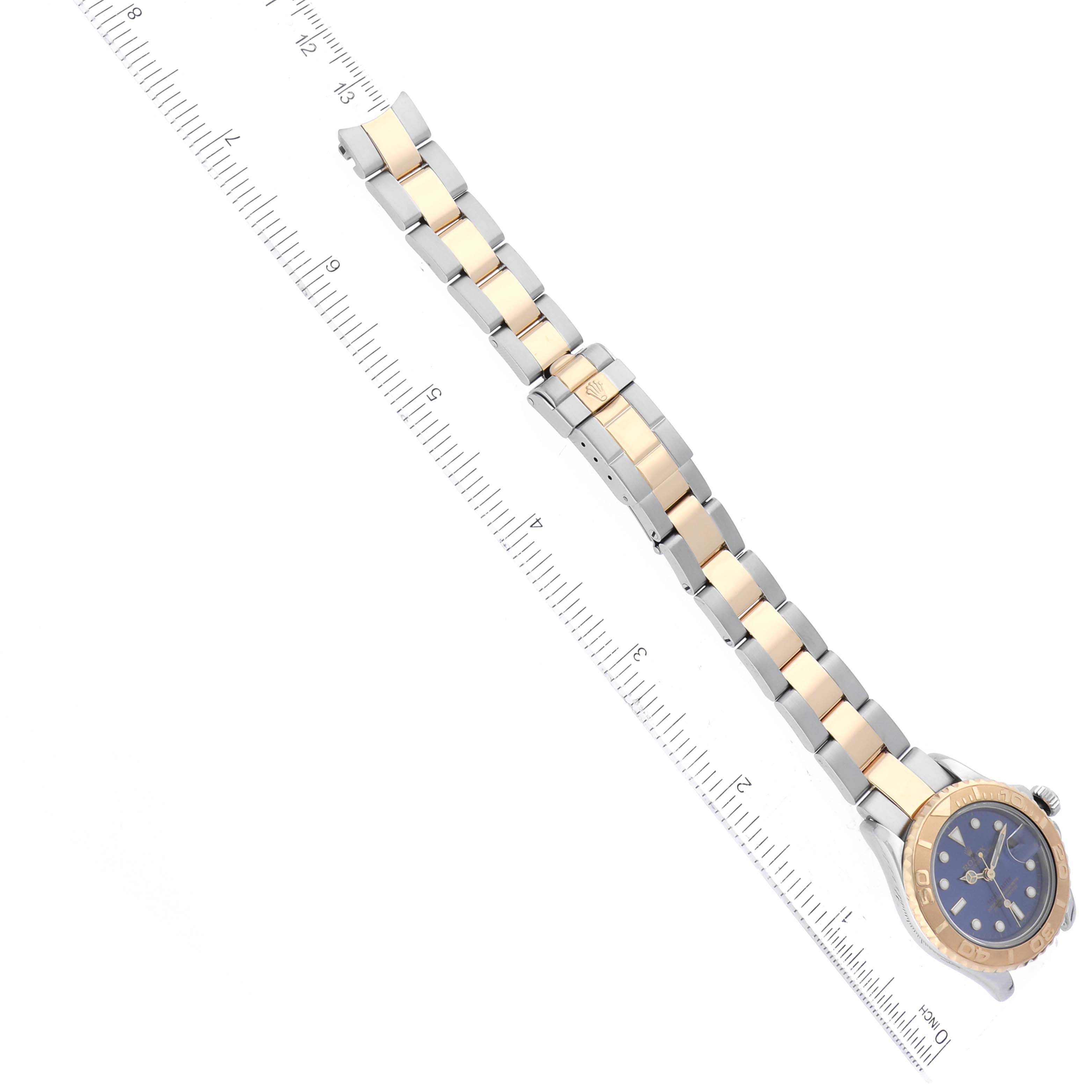 The image shows the Rolex Yacht-Master 69623 Women's Steel and Gold (two tone) Blue Dial 69623 Women's Steel and Gold (two tone) Blue Dial watch at an oblique angle, highlighting the bracelet, clasp, and watch face next to a ruler.