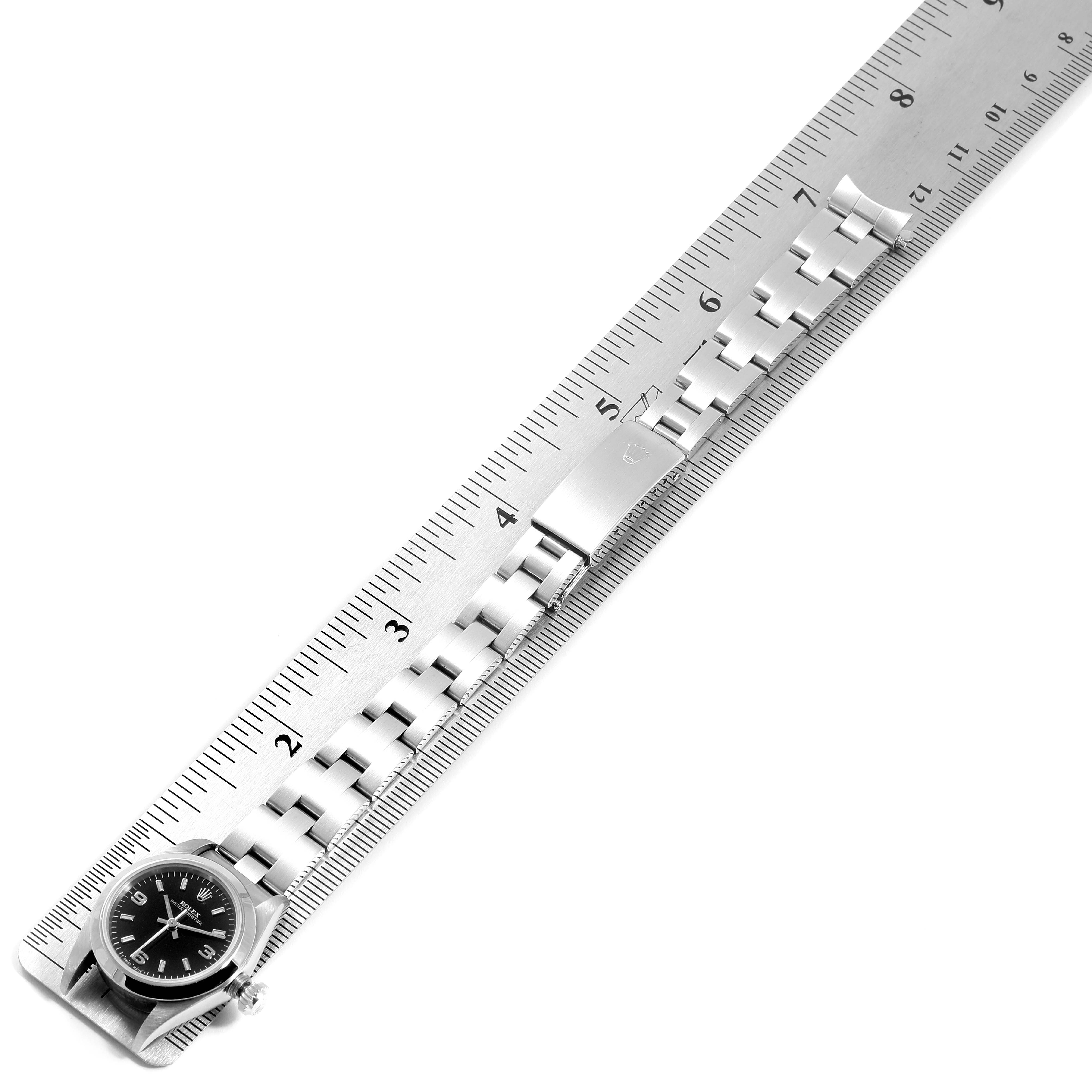 The image shows a Rolex Oyster Perpetual 67180 Women
s Stainless Steel Black Dial 67180 Women
s Stainless Steel Black Dial watch and band placed alongside a ruler, viewed from a top-down angle.