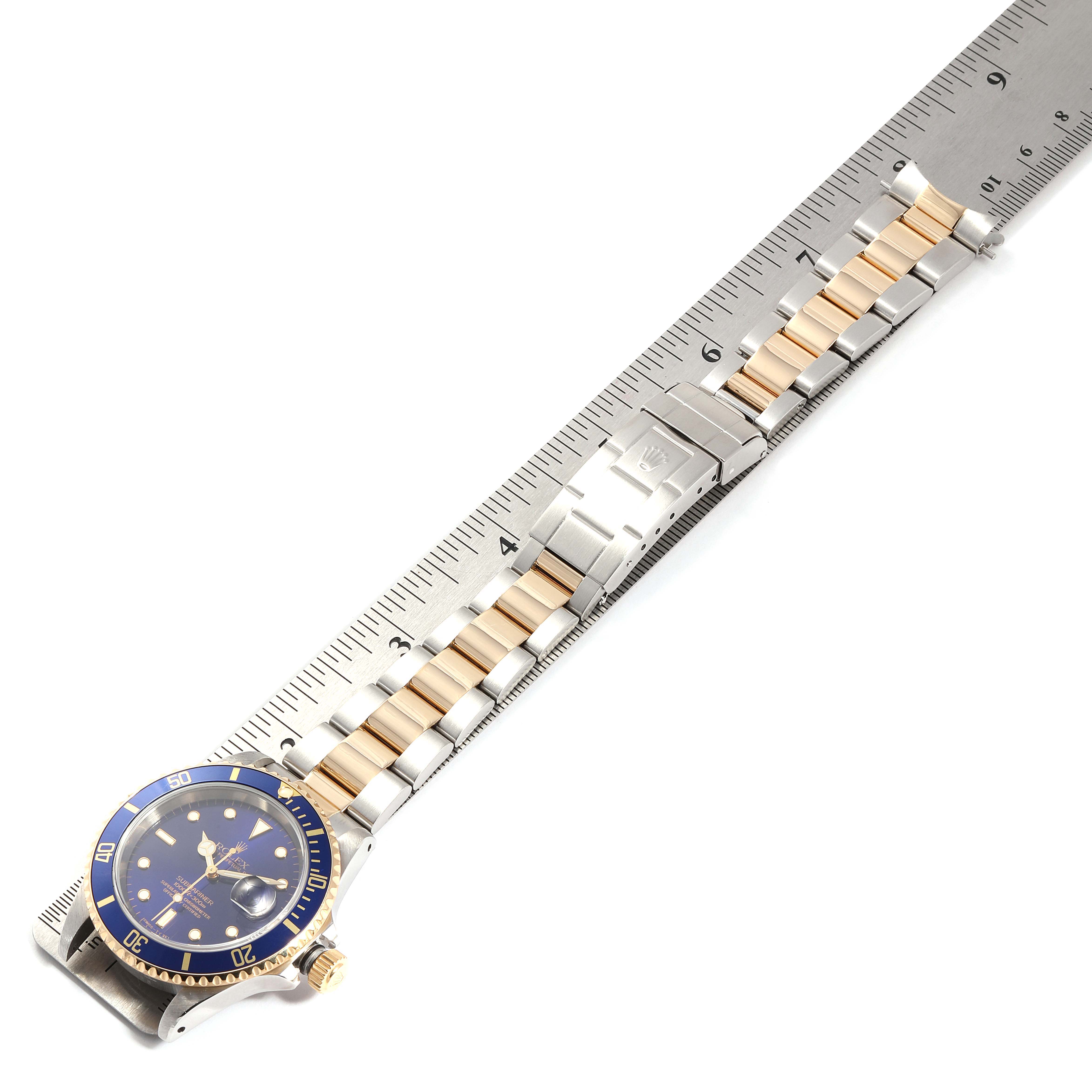 The image shows a Rolex Submariner 16613 Men
s Steel and Gold (two tone) Blue Dial 16613 Men
s Steel and Gold (two tone) Blue Dial watch laid flat, showcasing the face, bezel, and two-tone bracelet alongside a measuring ruler.