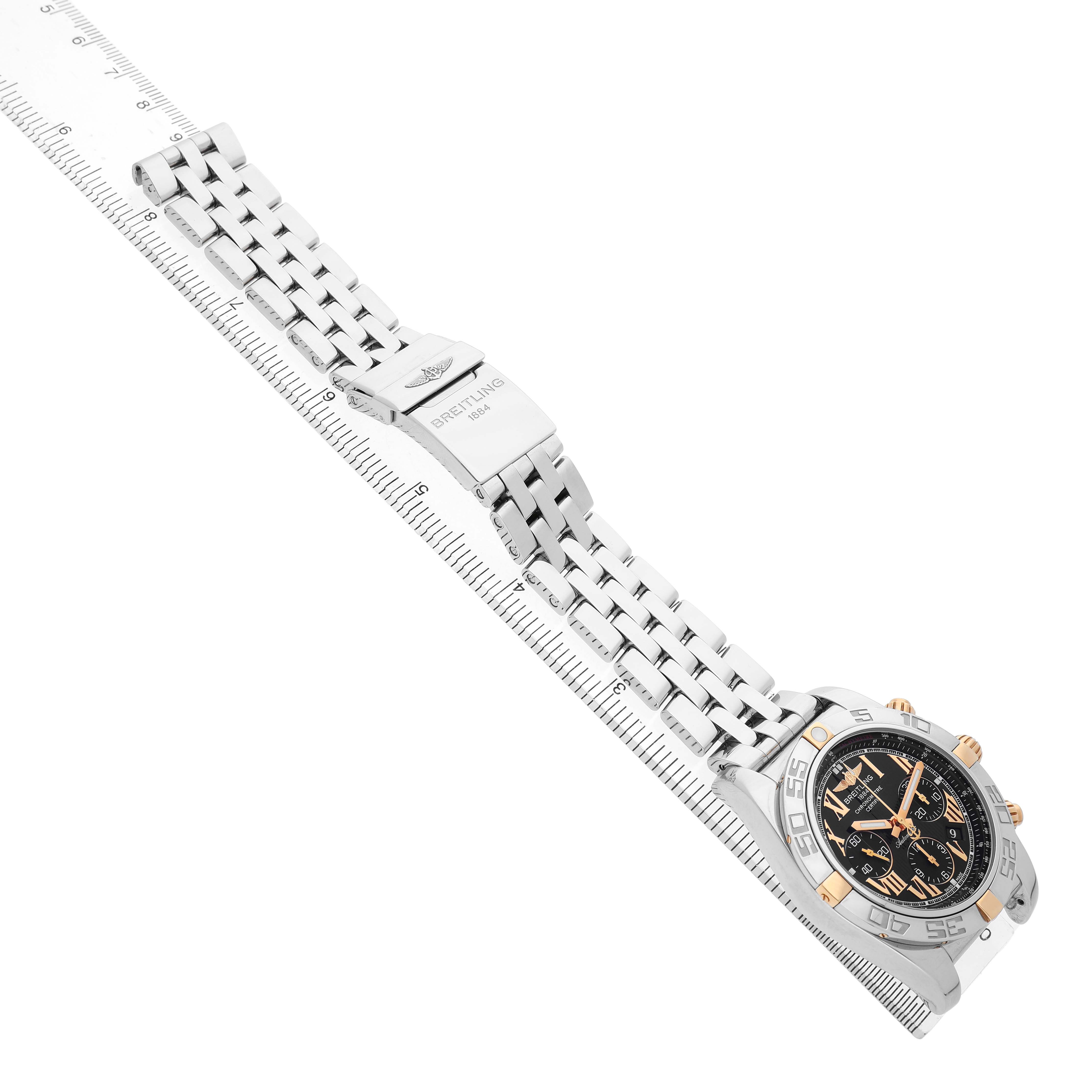 The Breitling Chronomat IB0110 Men's Steel and Gold (two tone) Black Dial watch is shown at an angled side view, displaying its face and metal bracelet with a clasp.