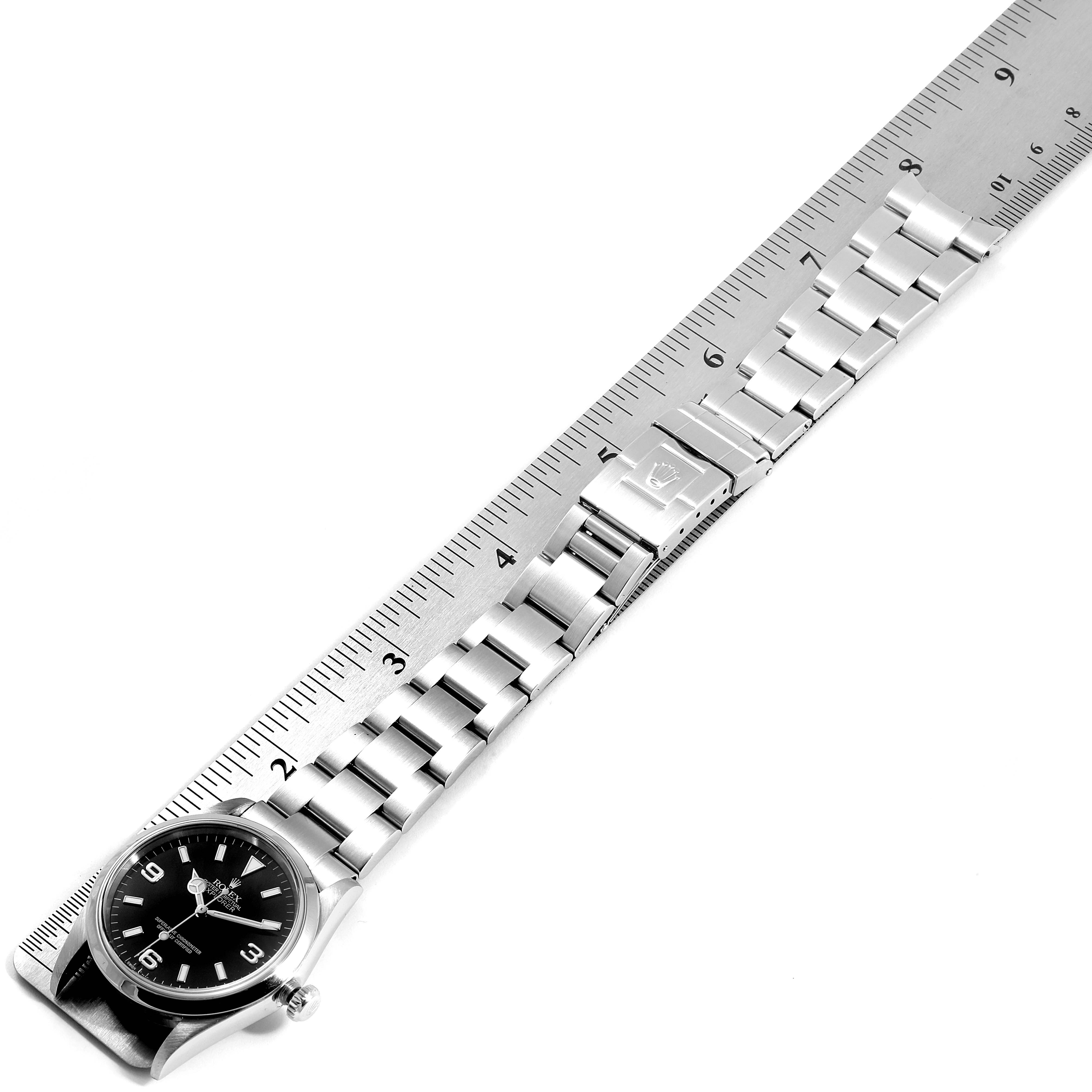 The image shows an overhead view of a Rolex Explorer 114270 Men
s Stainless Steel Black Dial 114270 Men
s Stainless Steel Black Dial watch, featuring its stainless steel band and clasp, next to a ruler for scale.
