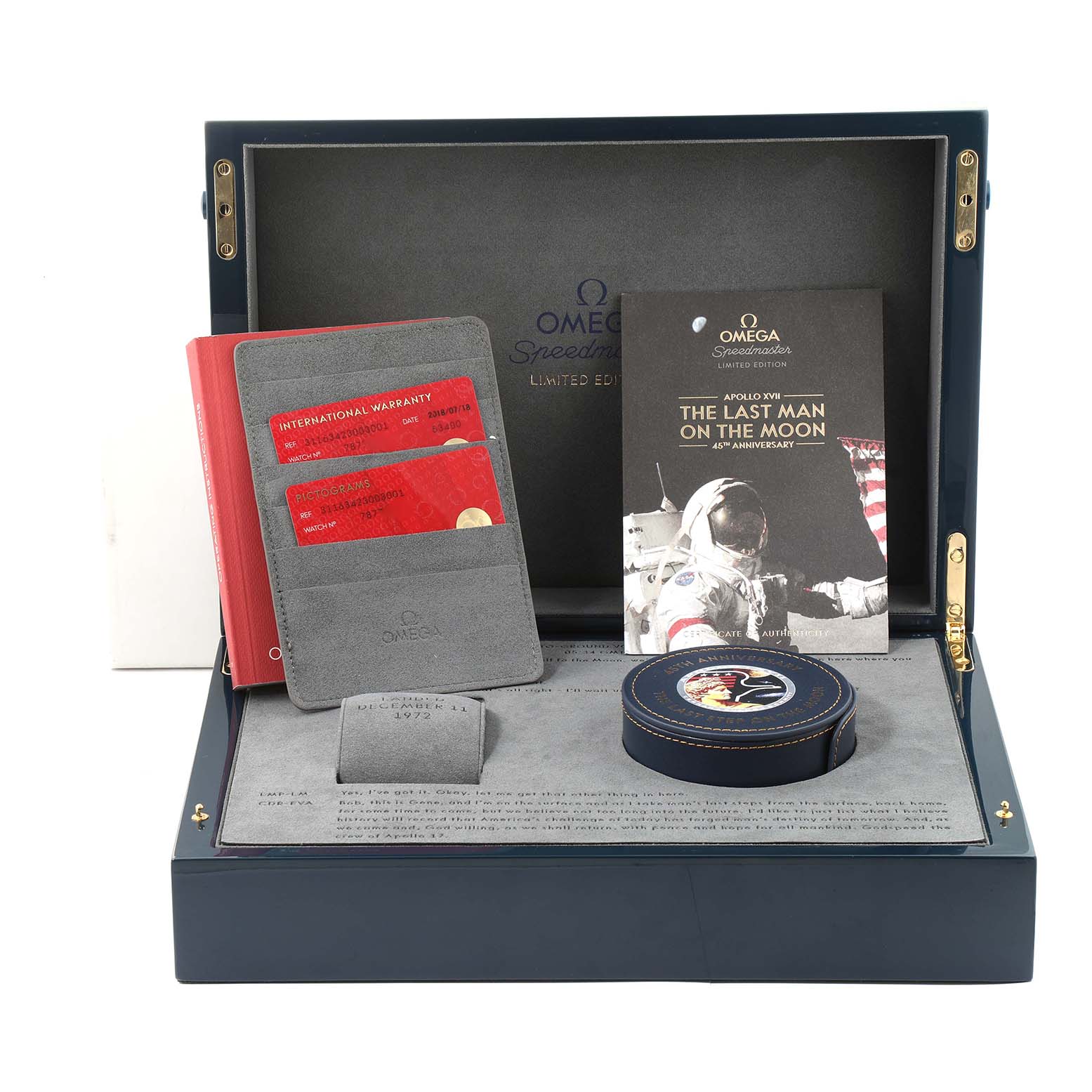 The image shows the packaging box of an Omega Speedmaster 311.63.42.30.03.001 Men's Yellow Gold Blue Dial 311.63.42.30.03.001 Men's Yellow Gold Blue Dial, containing a booklet, international warranty cards, and a commemorative patch.