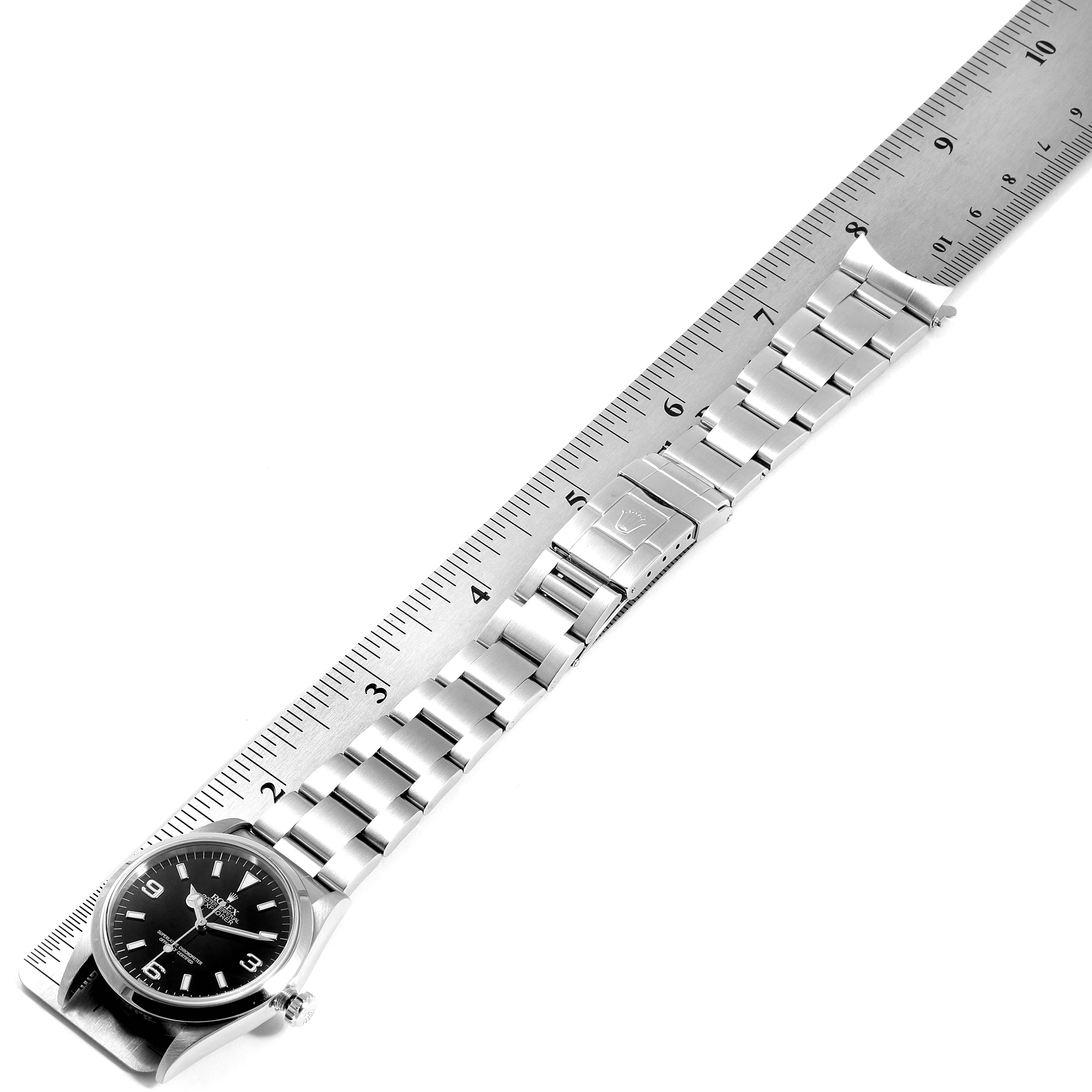 The Rolex Explorer 114270 Men
s Stainless Steel Black Dial 114270 Men
s Stainless Steel Black Dial watch is displayed flat with a metal ruler, showcasing the watch face and bracelet.