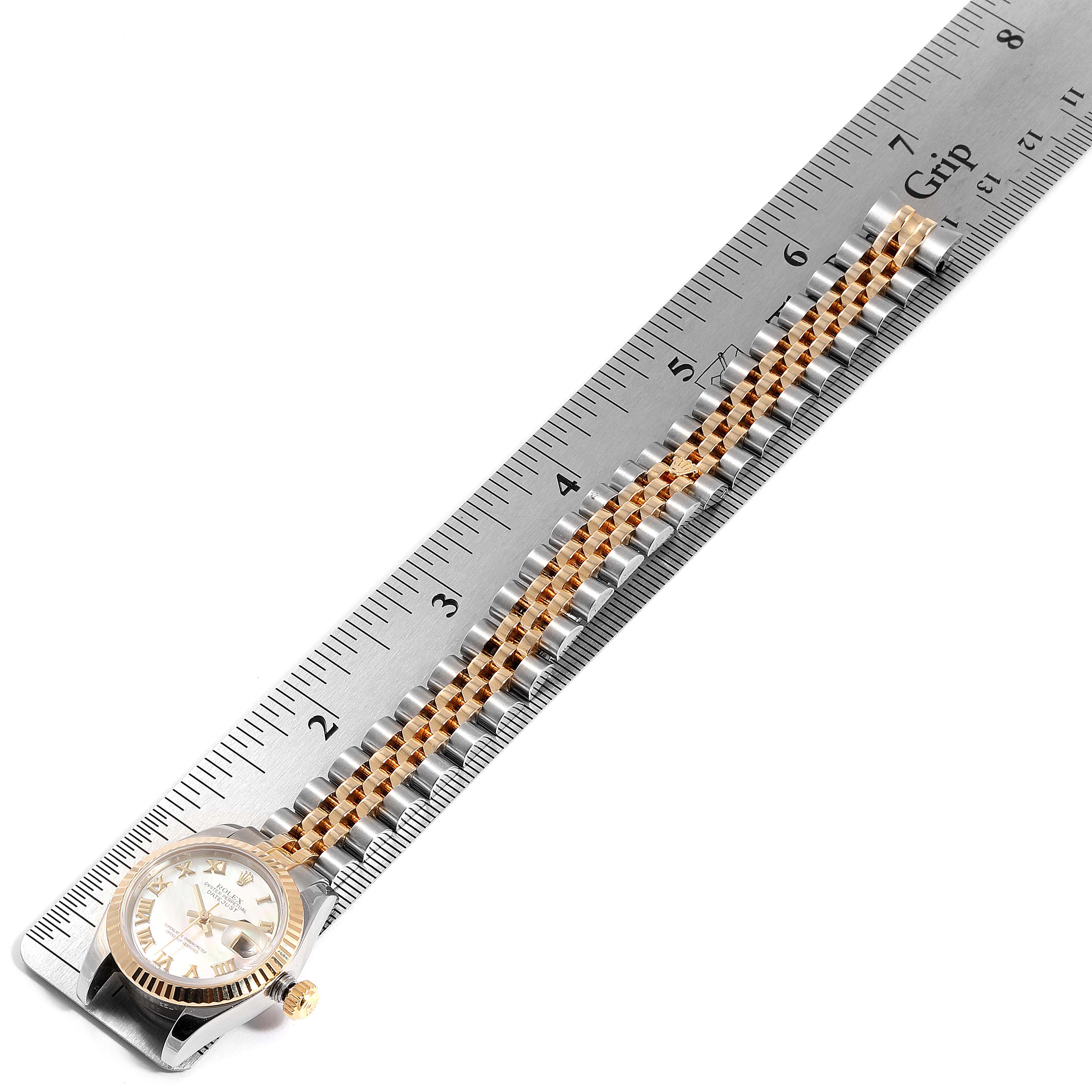 The Rolex Datejust 179173 Women's Steel and Gold (two tone) Mother of Pearl Dial 179173 Women's Steel and Gold (two tone) Mother of Pearl Dial watch is shown from a top-down angle, displaying the face, bezel, and fully extended bracelet alongside a ruler.