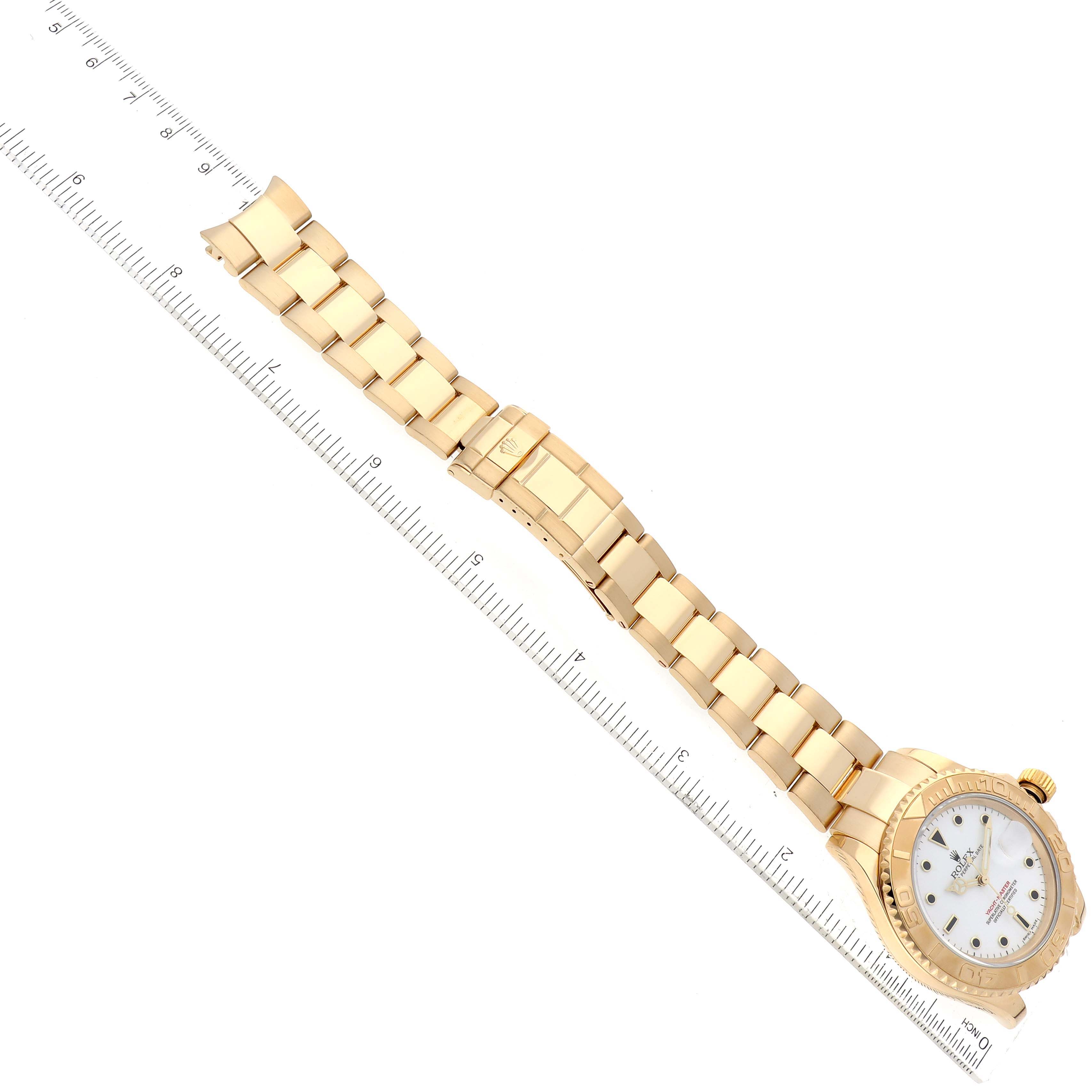 The image shows a top-down view of a Rolex Yacht-Master 16628 Men's Yellow Gold White Dial 16628 Men's Yellow Gold White Dial watch, displaying its gold band, dial, and bezel next to a ruler.