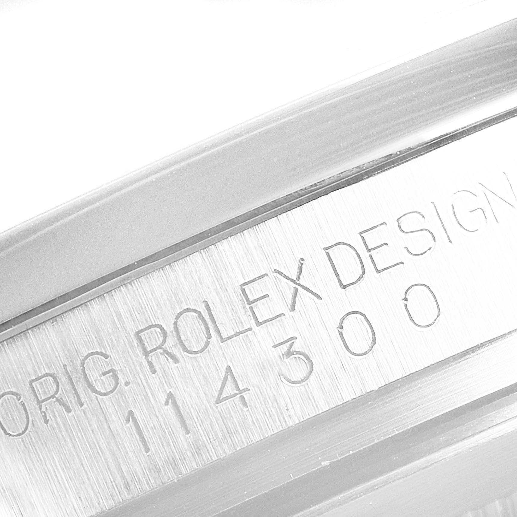 The image shows a close-up of the engraved text "ORIG. ROLEX DESIGN 114300" on the case of an Oyster Perpetual model by Rolex.