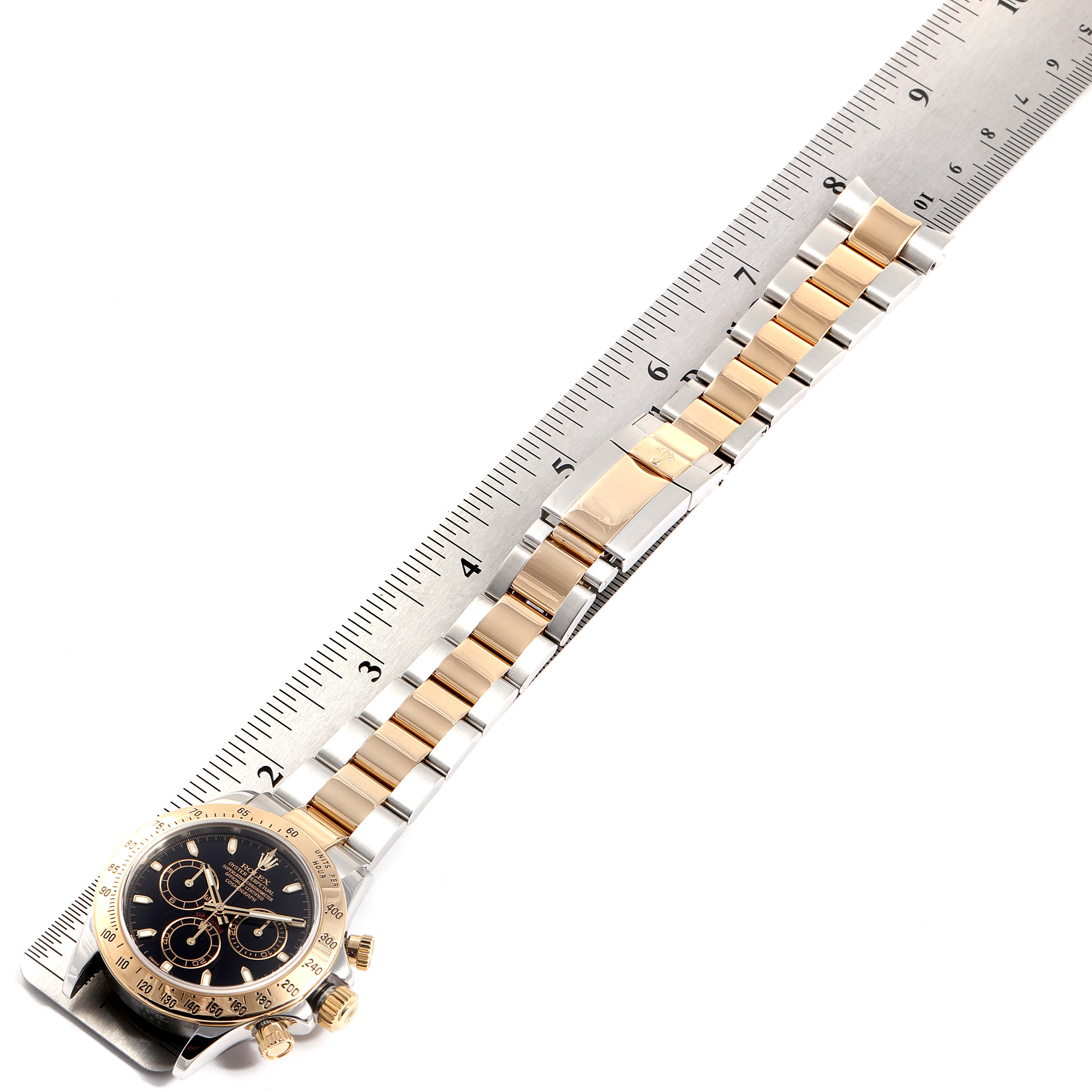 The image shows a Rolex Daytona 116523 Men
s Steel and Gold (two tone) Black Dial 116523 Men
s Steel and Gold (two tone) Black Dial watch with a black dial and gold accents, laid flat next to a ruler.