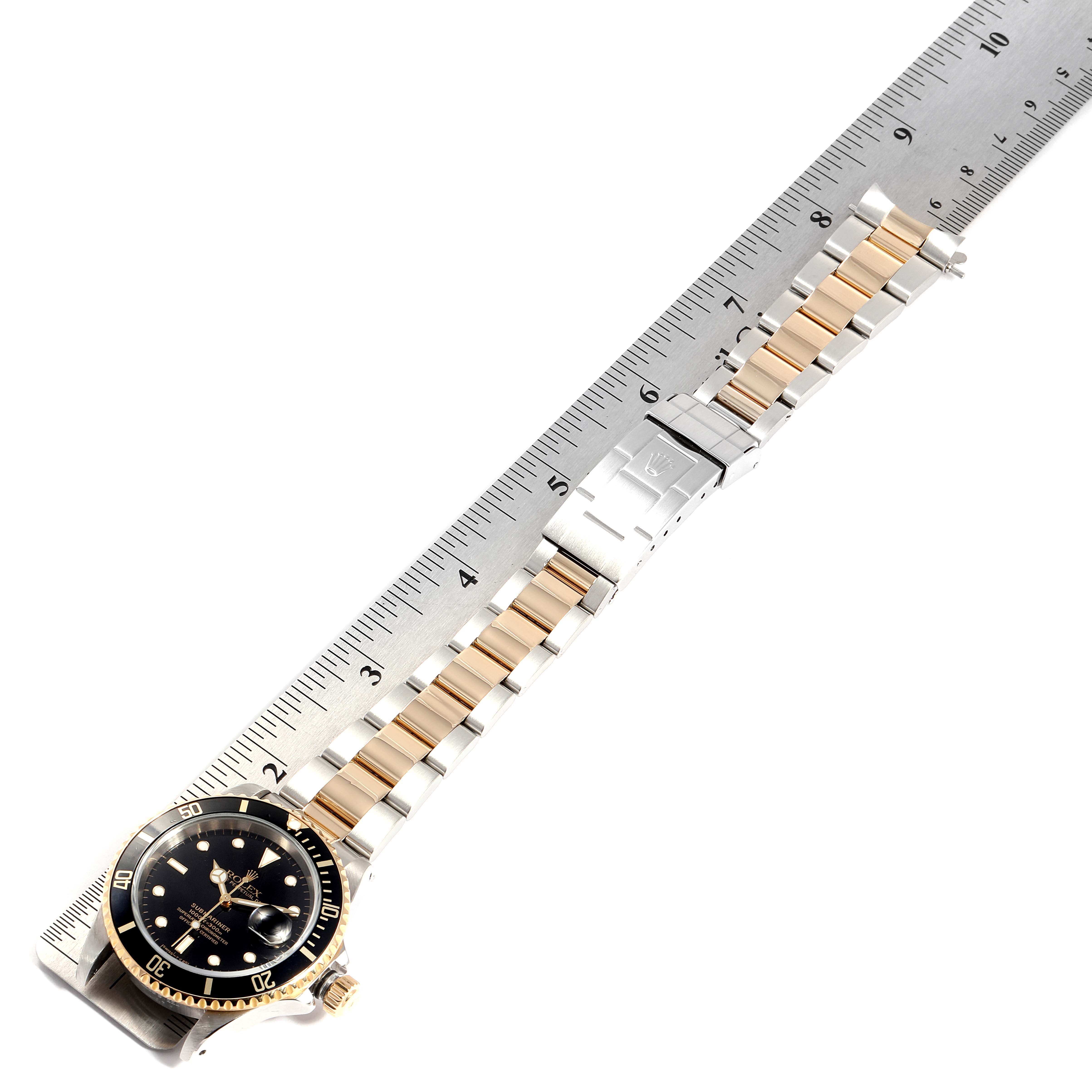 The image shows a Rolex Submariner 16613 Men
s Steel and Gold (two tone) Black Dial 16613 Men
s Steel and Gold (two tone) Black Dial watch, laid flat alongside a ruler, detailing the face, bezel, bracelet, and clasp.