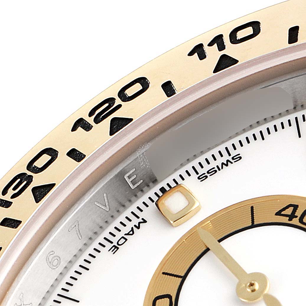 This close-up image of a Rolex Daytona 116503 Men
s Steel and Gold (two tone) White Dial 116503 Men
s Steel and Gold (two tone) White Dial watch shows the bezel with tachymeter markings and part of the dial with a hand and marker.