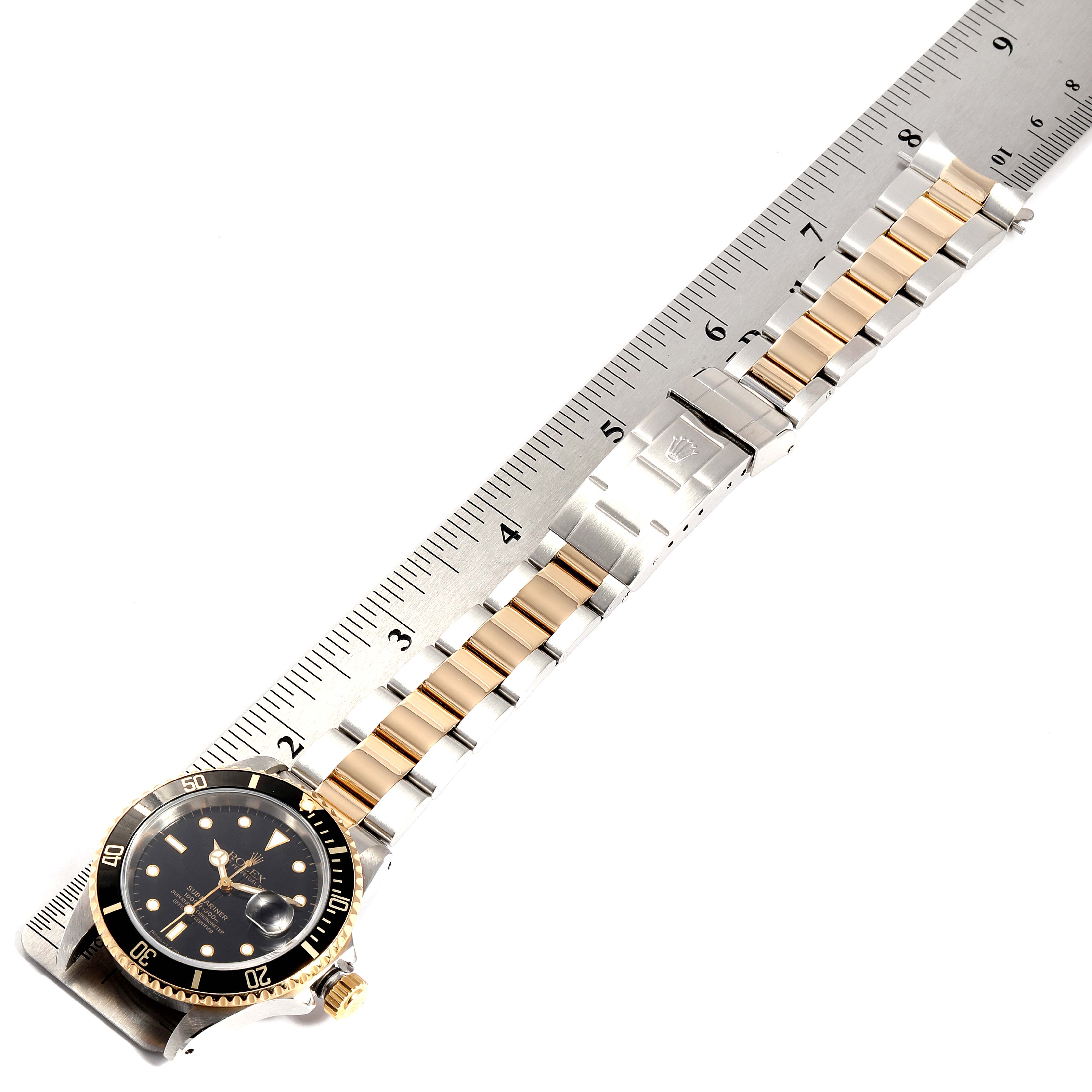 The image shows a Rolex Submariner 16613 Men
s Steel and Gold (two tone) Black Dial 16613 Men
s Steel and Gold (two tone) Black Dial watch viewed from above, with the watch band also visible along with a ruler for measurement.