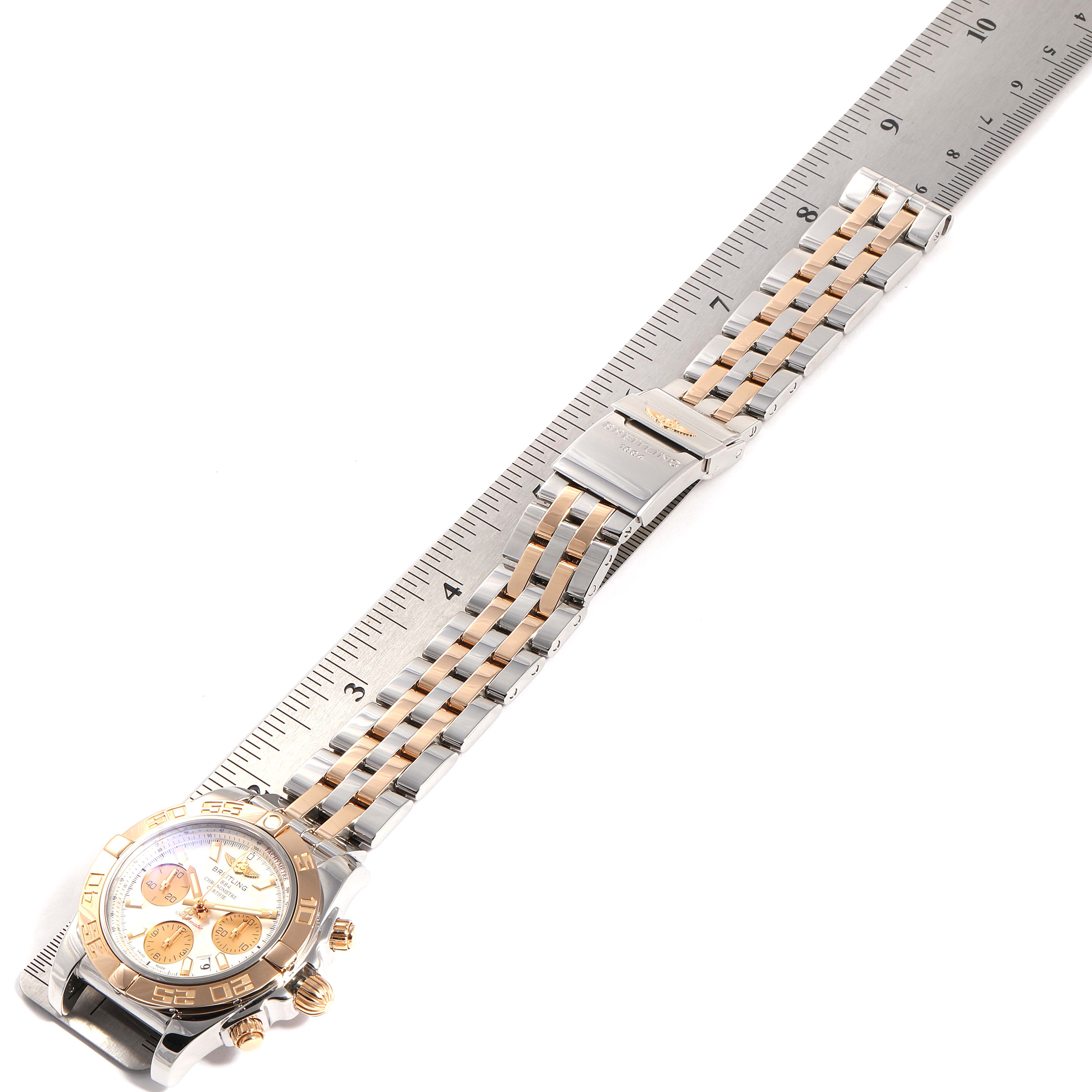 The Breitling Chronomat CB0140Y2/A743-378C Men's Steel and Gold (two tone) Silver Dial CB0140Y2/A743-378C Men's Steel and Gold (two tone) Silver Dial watch is shown from a top-down angle, displaying its face, bezel, and metal strap alongside a ruler.