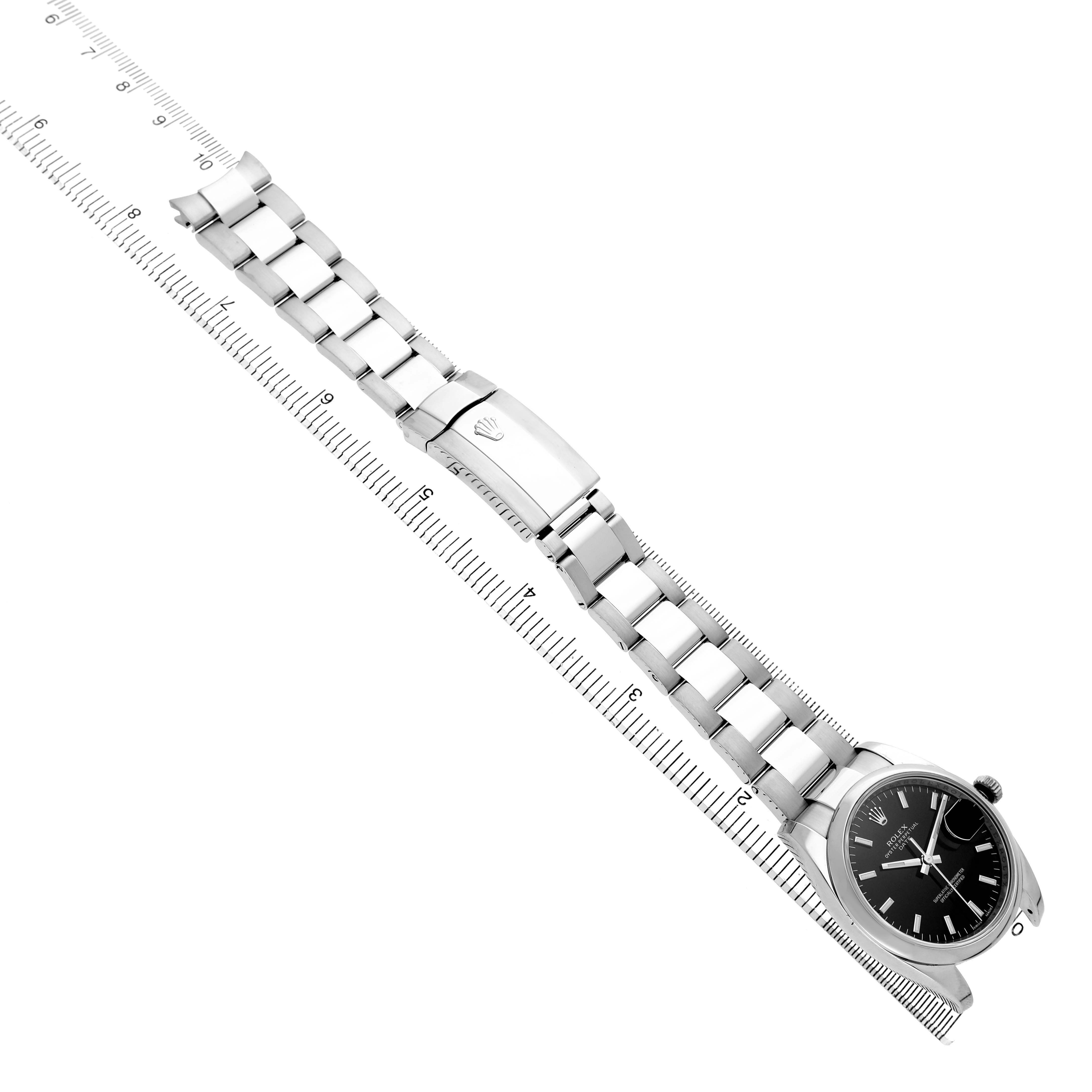 The Rolex Date 115200 Men's Stainless Steel Black Dial watch is shown at an angle displaying the bracelet, clasp, and partial view of the dial alongside a ruler.