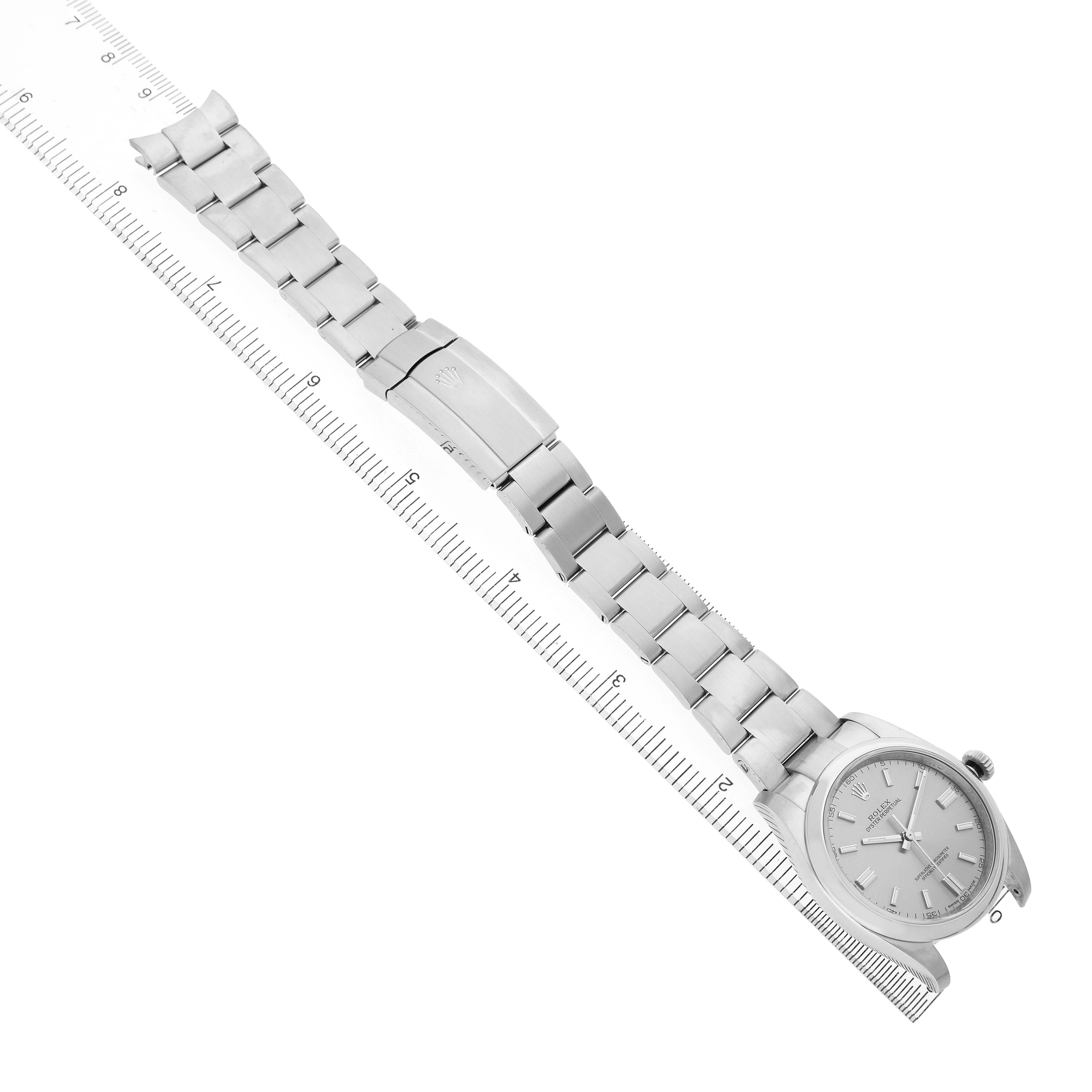 The Rolex Oyster Perpetual 116000 Men's Stainless Steel Silver Dial watch is shown from an overhead angle, displaying the full bracelet, clasp, and face beside a ruler.