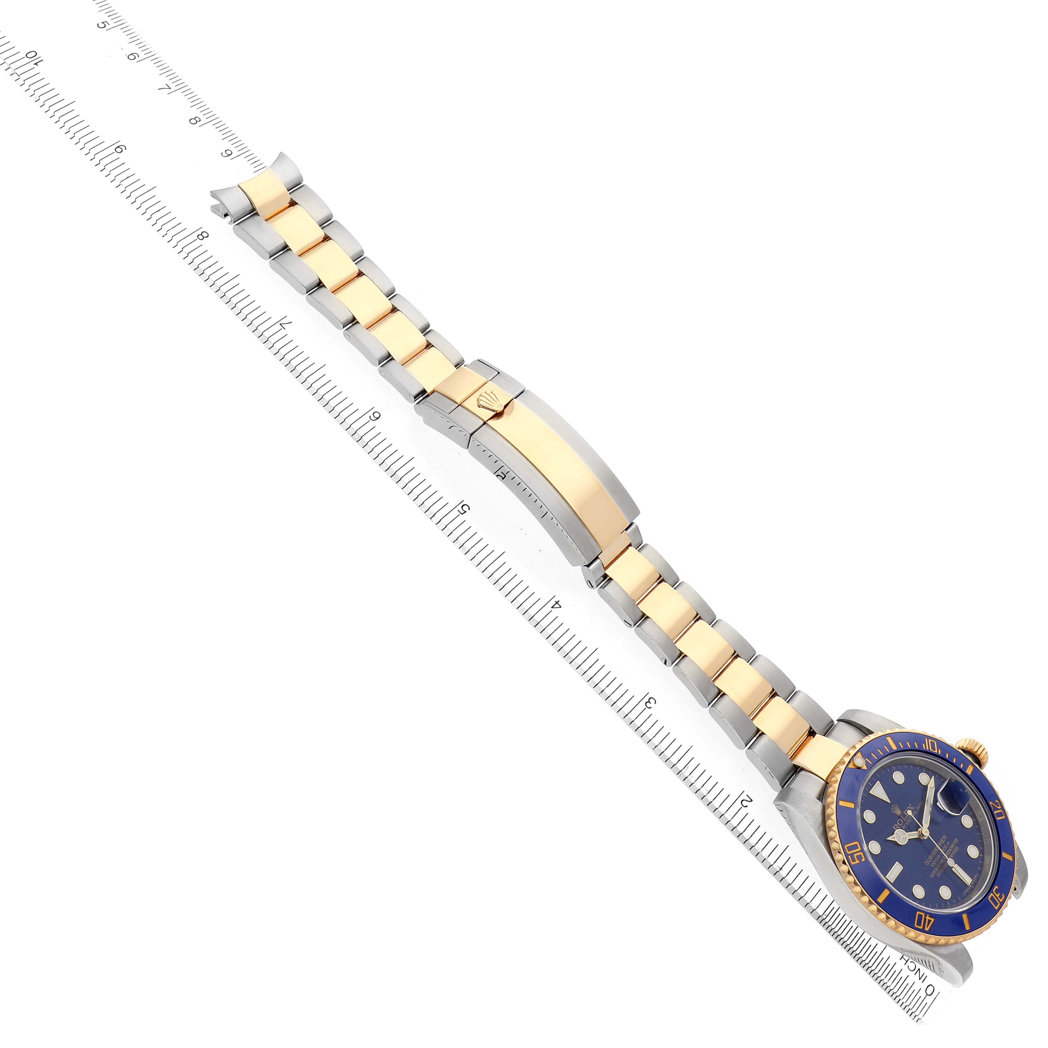 The image shows a Rolex Submariner 116613 Men
s Steel and Gold (two tone) Blue Dial watch at a diagonal angle with the face, bracelet, and clasp visible, alongside a ruler.
