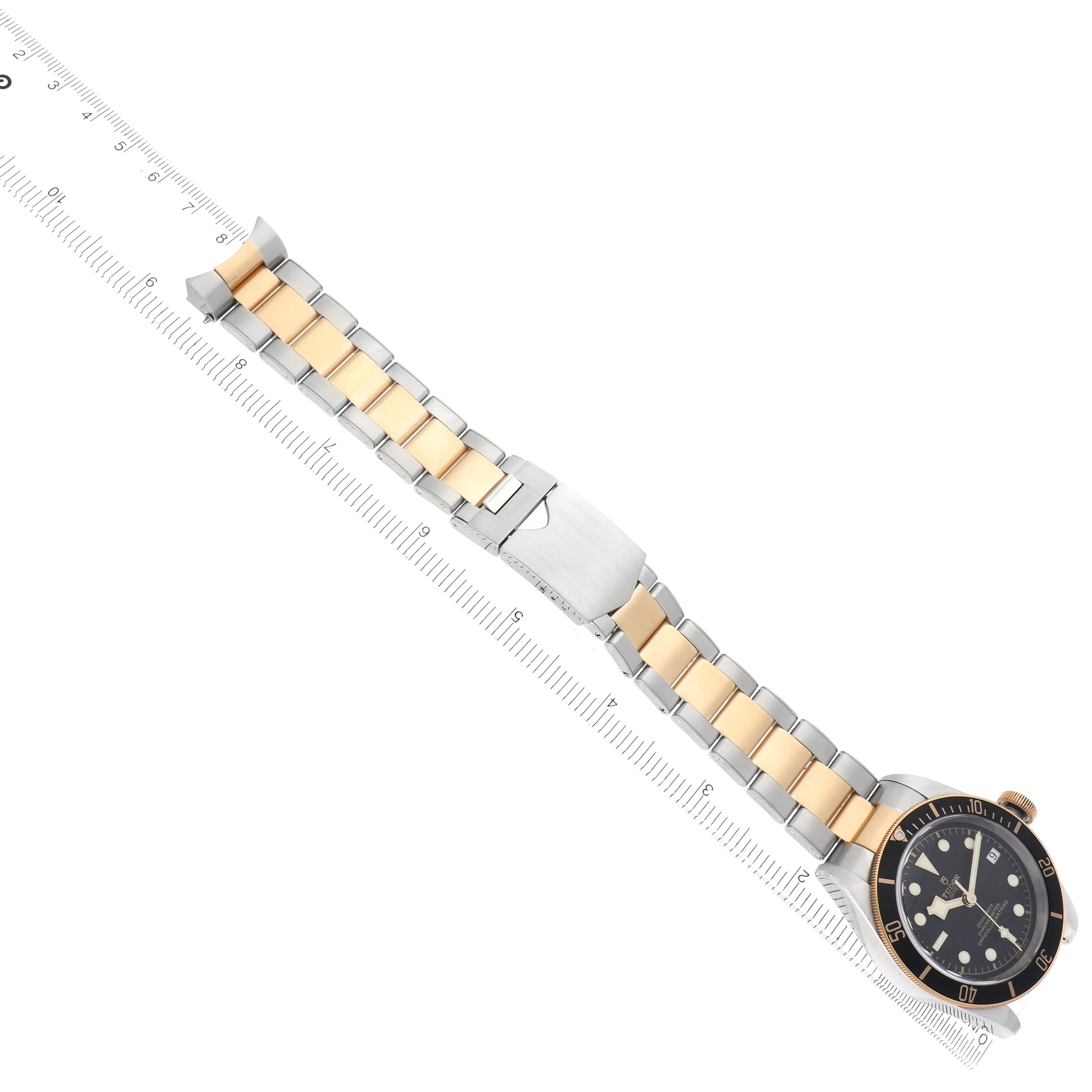The Tudor Heritage Black Bay 79733 Men
s Steel and Gold (two tone) Black Dial watch is shown at an angle highlighting the bracelet, clasp, and face alongside a ruler.