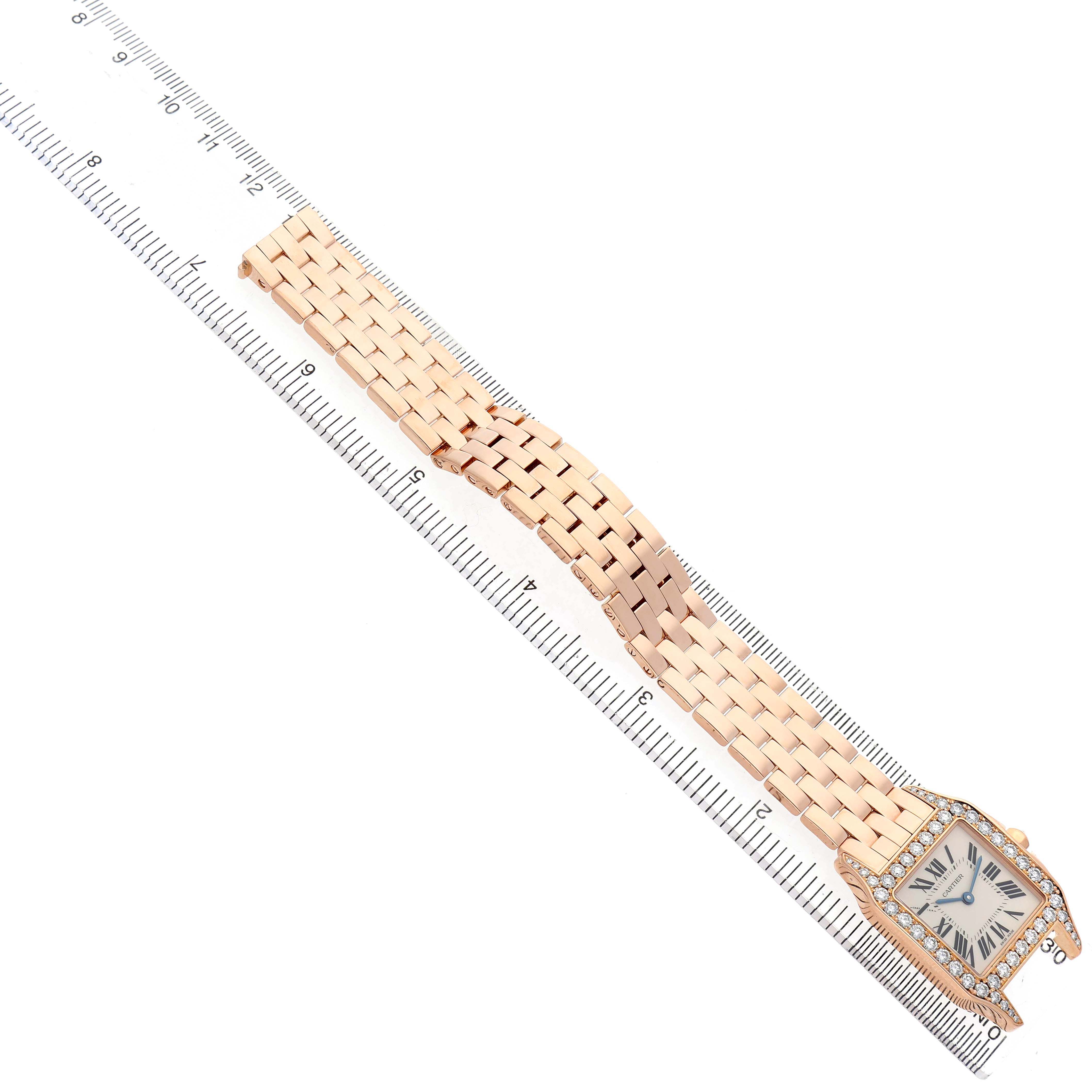 The Cartier Santos Demoiselle WF9007Z8 Women's Yellow Gold Silver Dial watch is shown top-down, highlighting the face, diamond bezel, and gold bracelet next to a ruler.