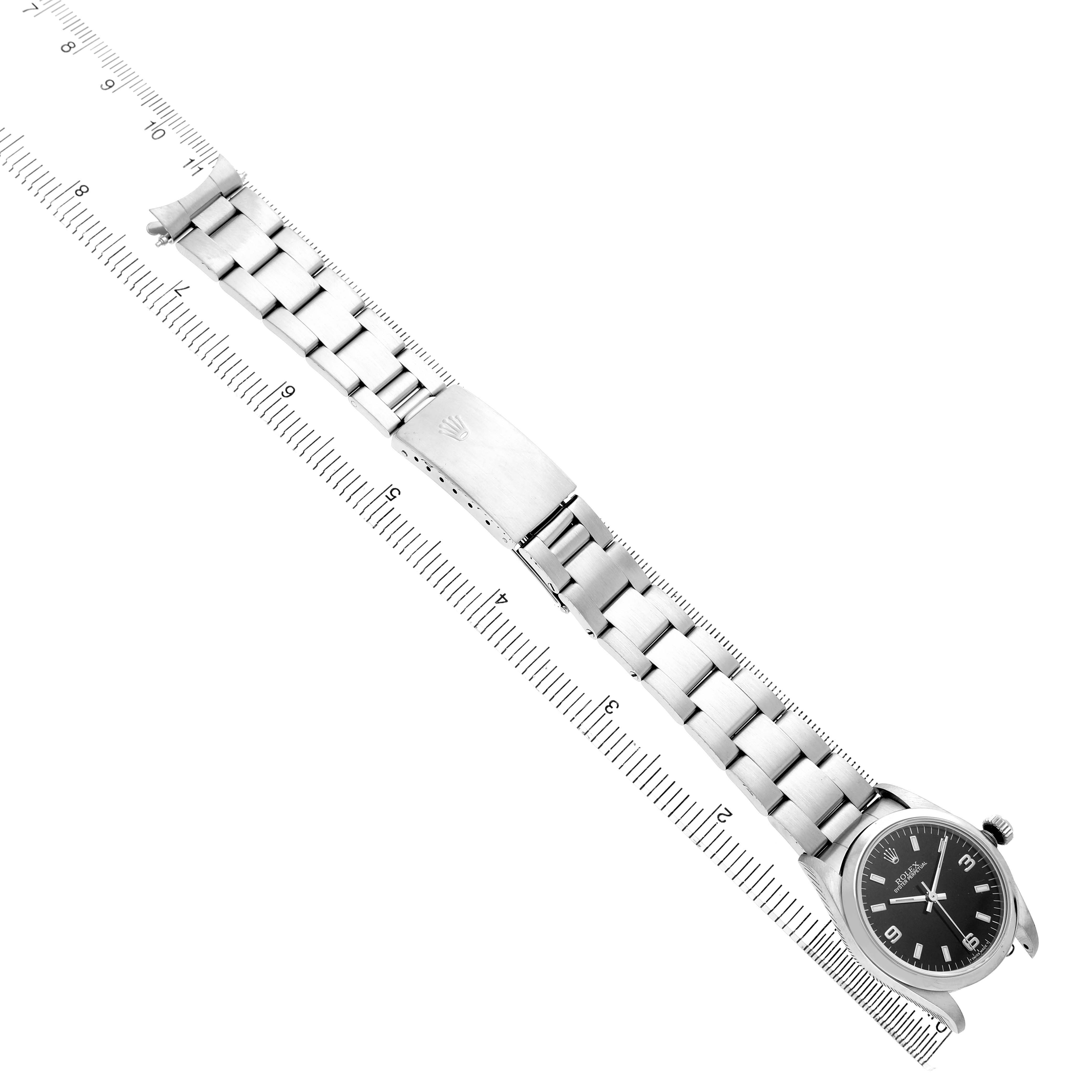 The Rolex Mid-Size 67480 Women's Stainless Steel Black Dial watch is displayed at an angle, showcasing its bracelet, clasp, and the face alongside a measuring ruler.