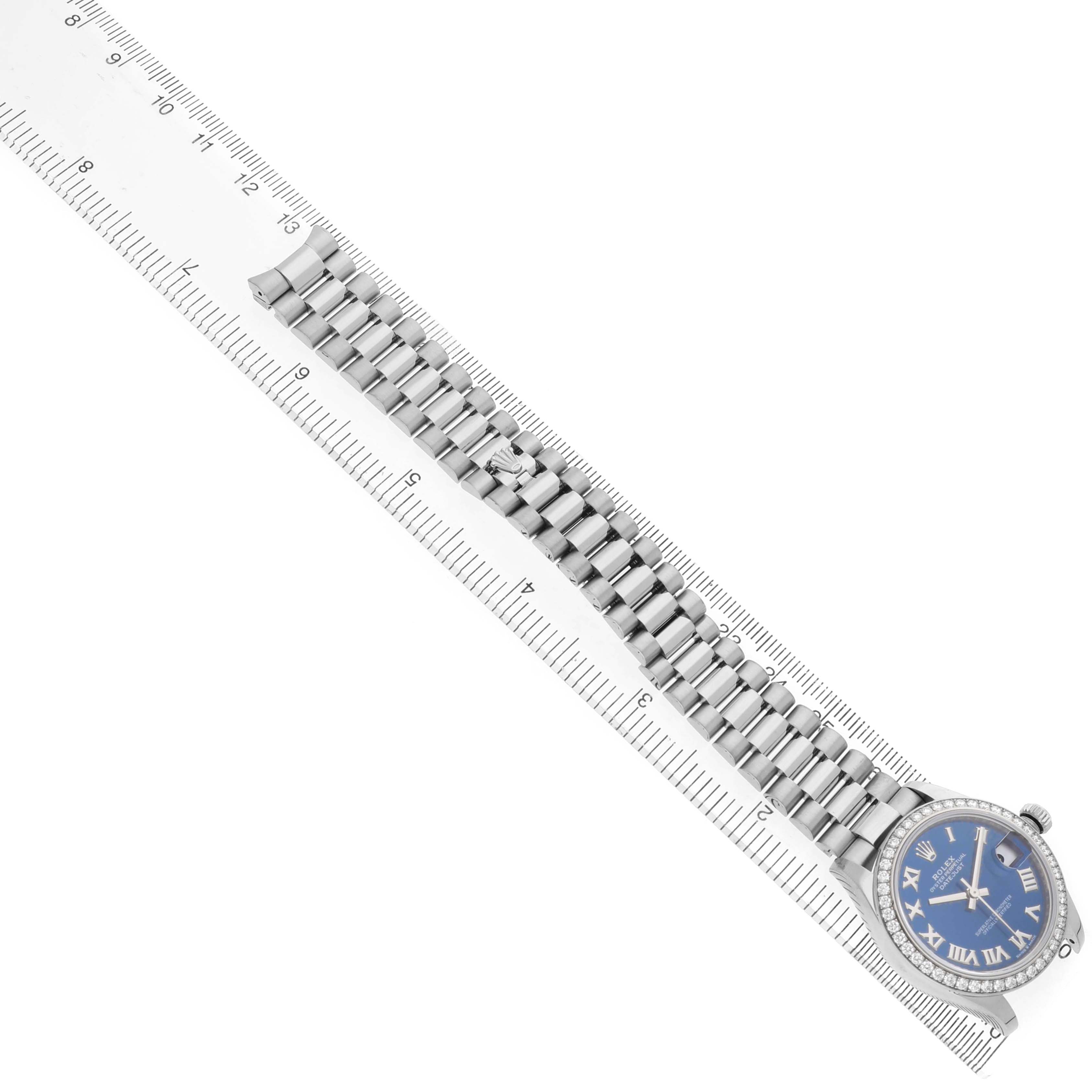 The Rolex President 278289 Women's White Gold Blue Dial model watch is shown lying flat alongside a ruler, displaying its bezel, bracelet, and face from a top-down angle.