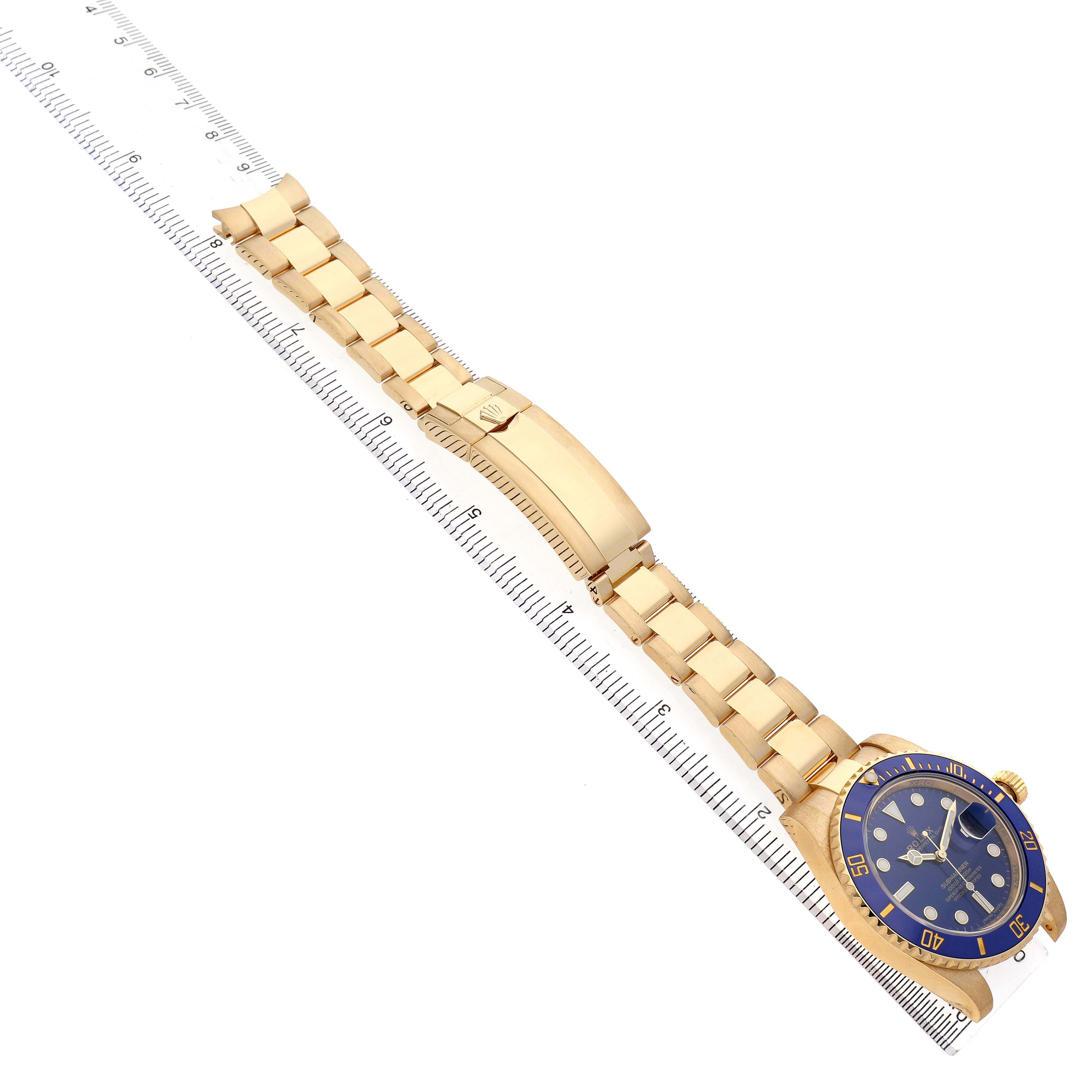 The Rolex Submariner 116618 Men's Yellow Gold Blue Dial watch is displayed diagonally, showing the bracelet, case, and clasp, aligned with a ruler.