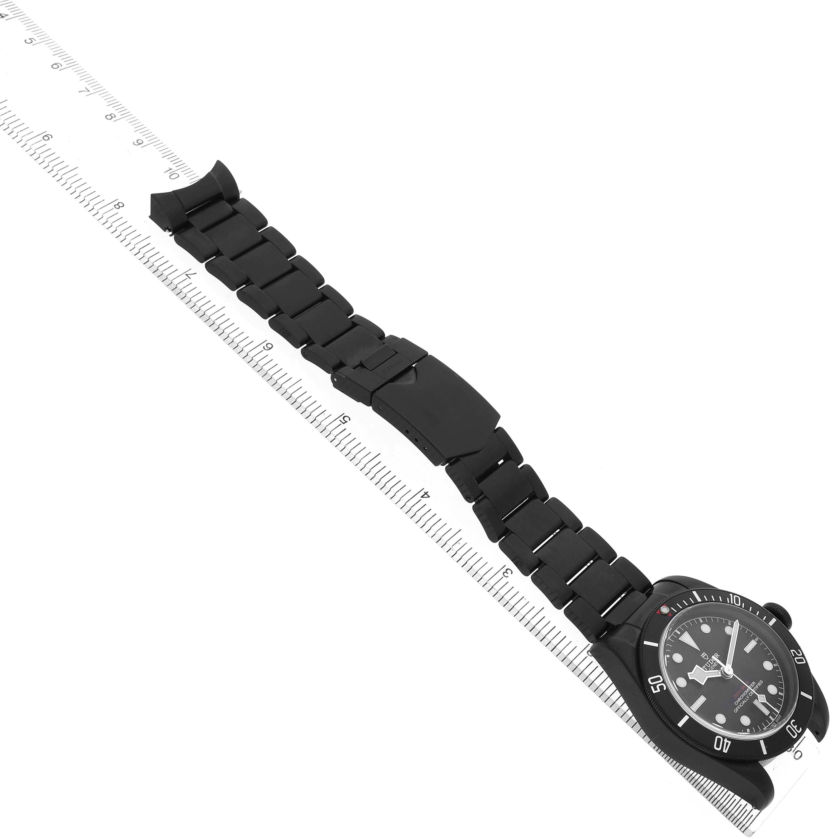 The Tudor Heritage Black Bay 79230DK Men's Stainless Steel Black Dial watch is shown from a top view, featuring the watch face and full bracelet alongside a ruler for scale.