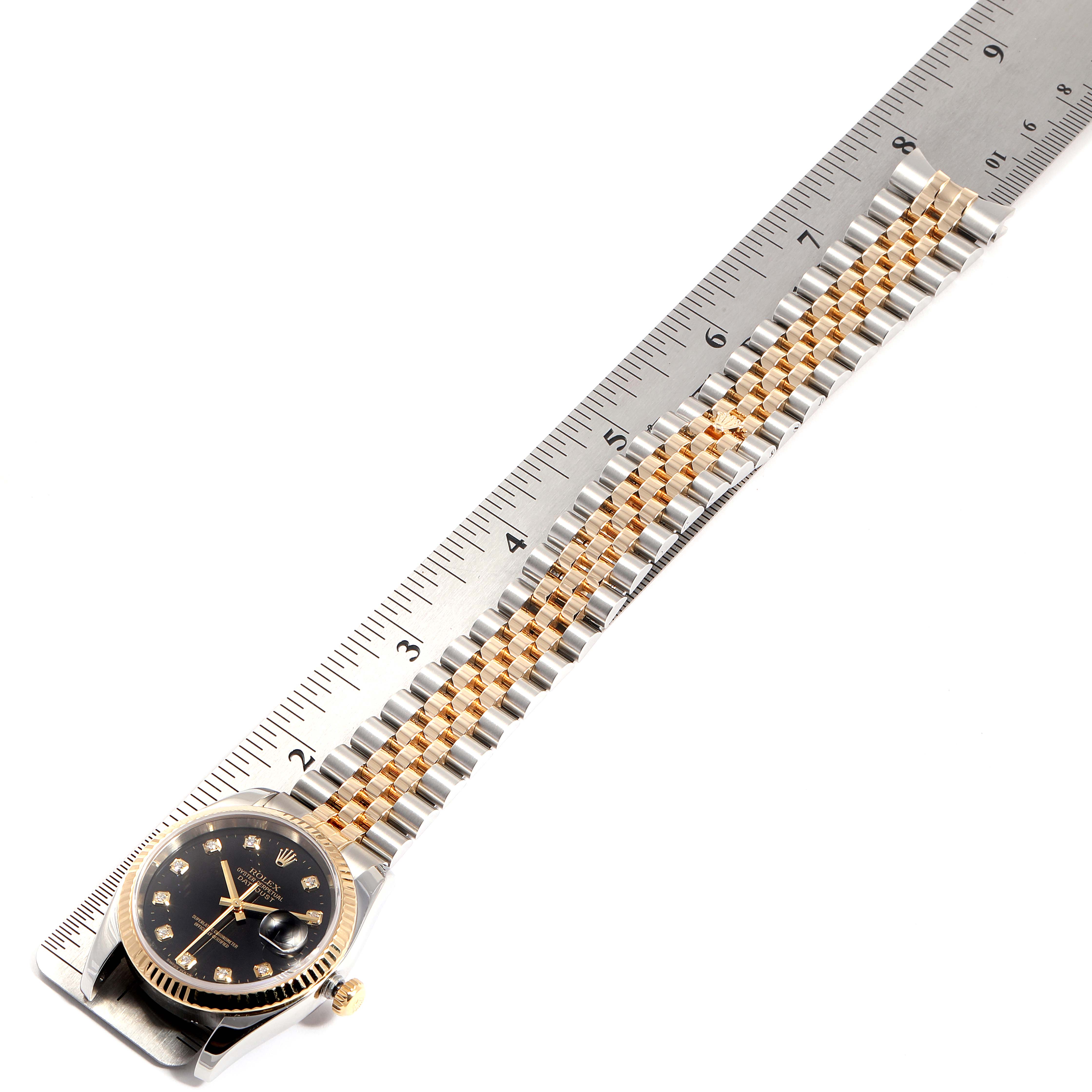 The Rolex Datejust 116233 Men
s Steel and Gold (two tone) Black Dial 116233 Men
s Steel and Gold (two tone) Black Dial watch is shown from a top view, highlighting its face, crown, and two-tone bracelet laid against a ruler.