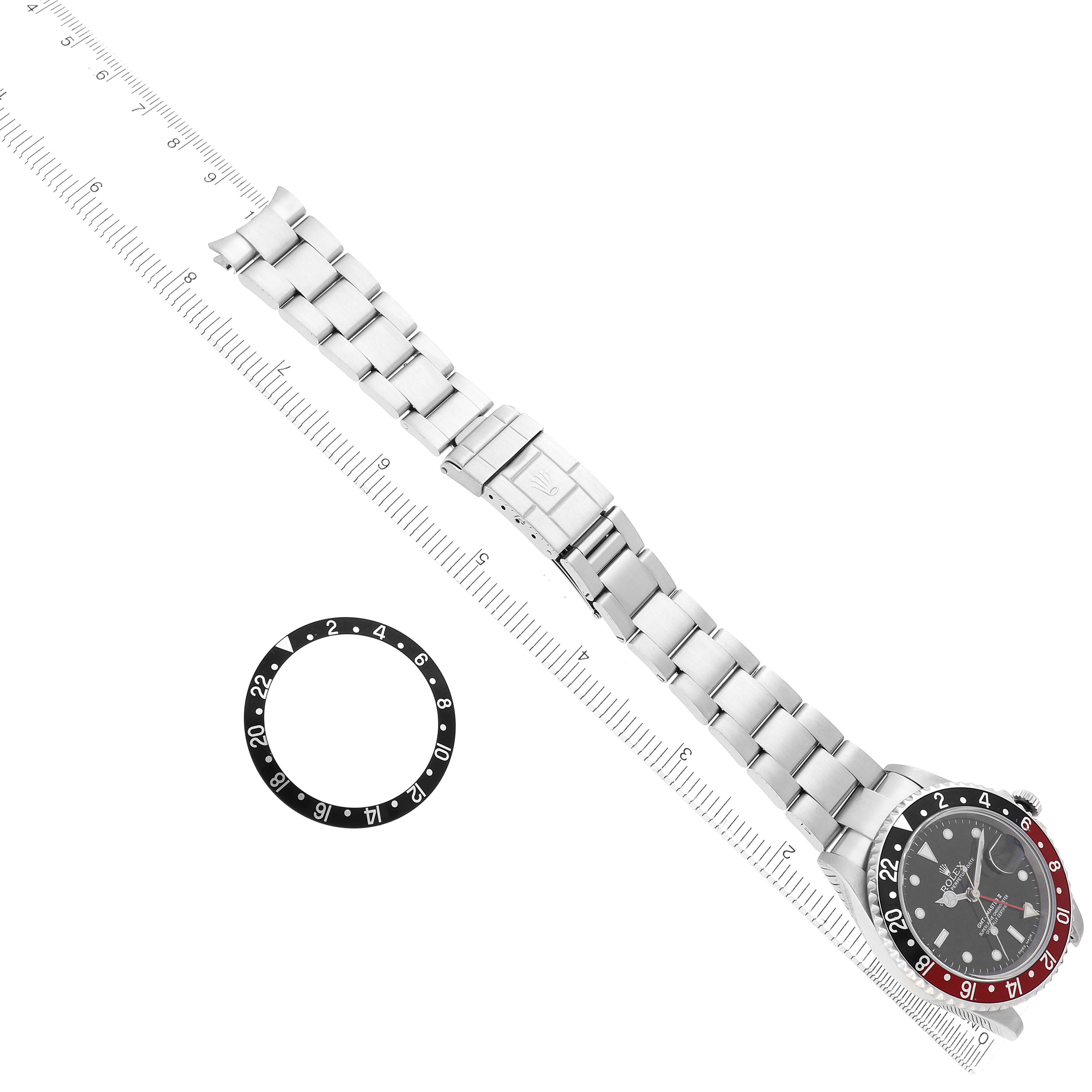 The Rolex GMT-Master 16710 Men
s Stainless Steel Black Dial watch is shown from above, with the bezel insert separated, alongside a bracelet and a ruler.