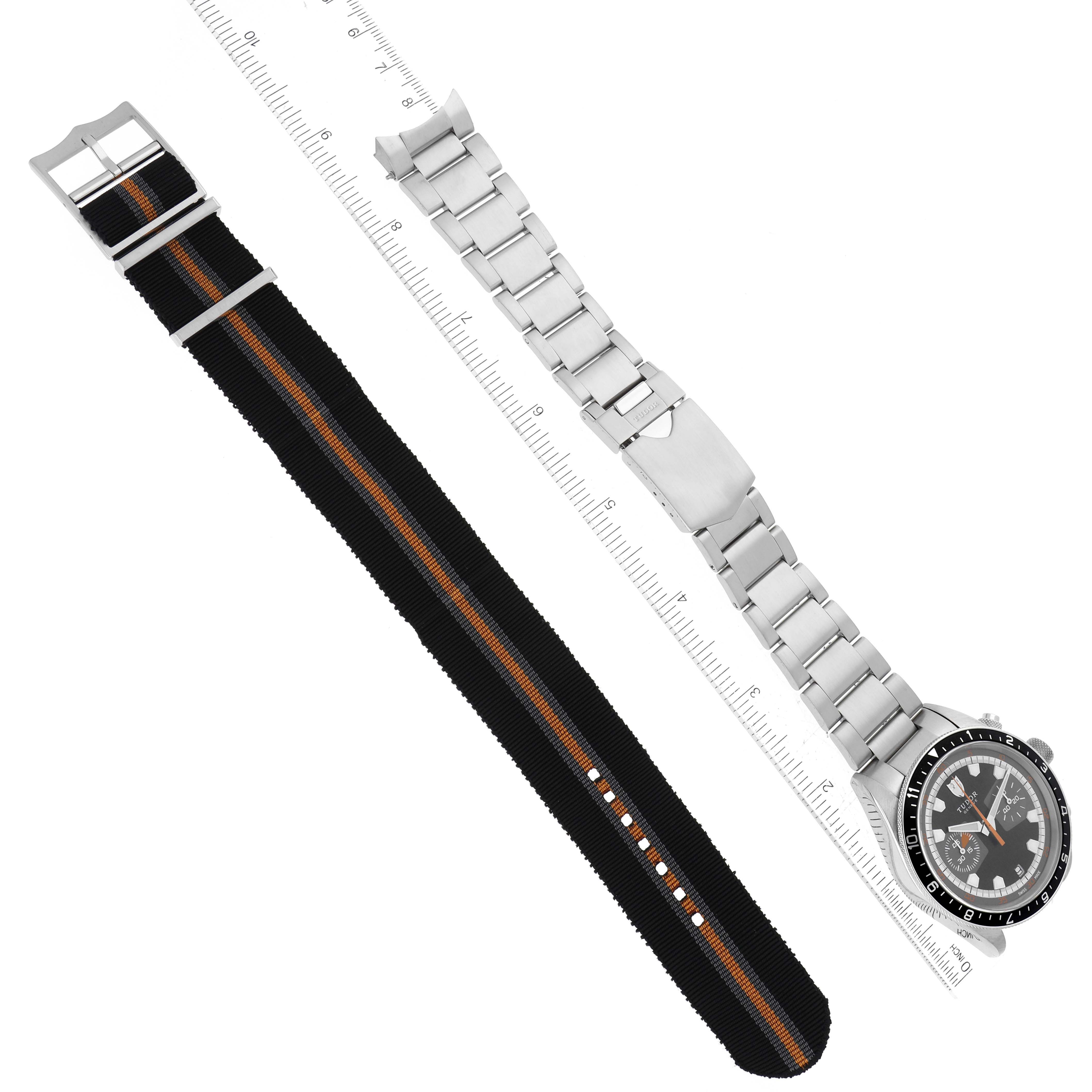This image shows a Tudor Heritage Chrono 70330N-95740 Men
s Stainless Steel Black Dial 70330N-95740 Men
s Stainless Steel Black Dial watch laid flat, featuring both its metal bracelet and fabric strap beside a ruler.