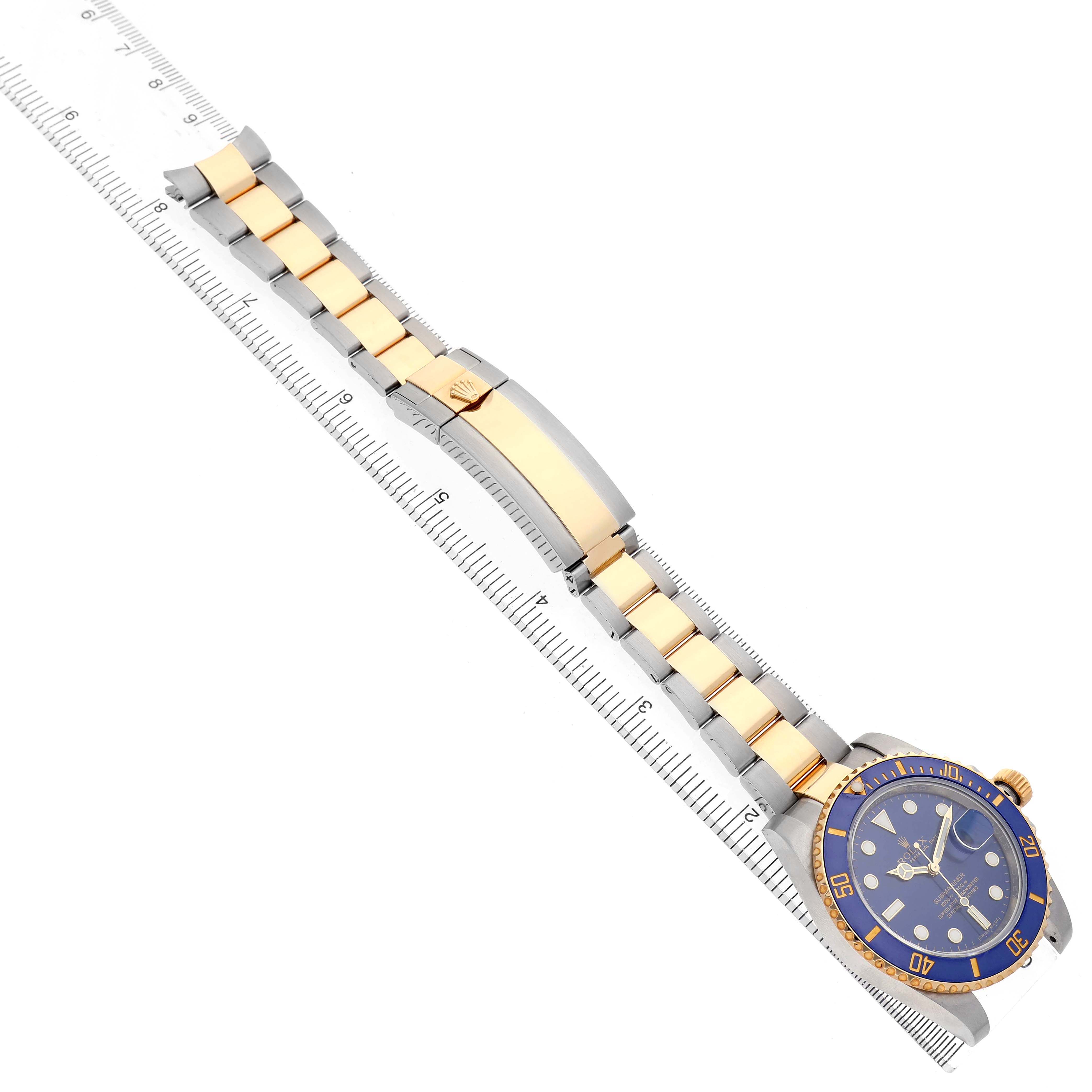 The Rolex Submariner 116613 Men's Steel and Gold (two tone) Blue Dial watch is shown diagonally with its full bracelet and clasp visible, alongside a ruler for scale.