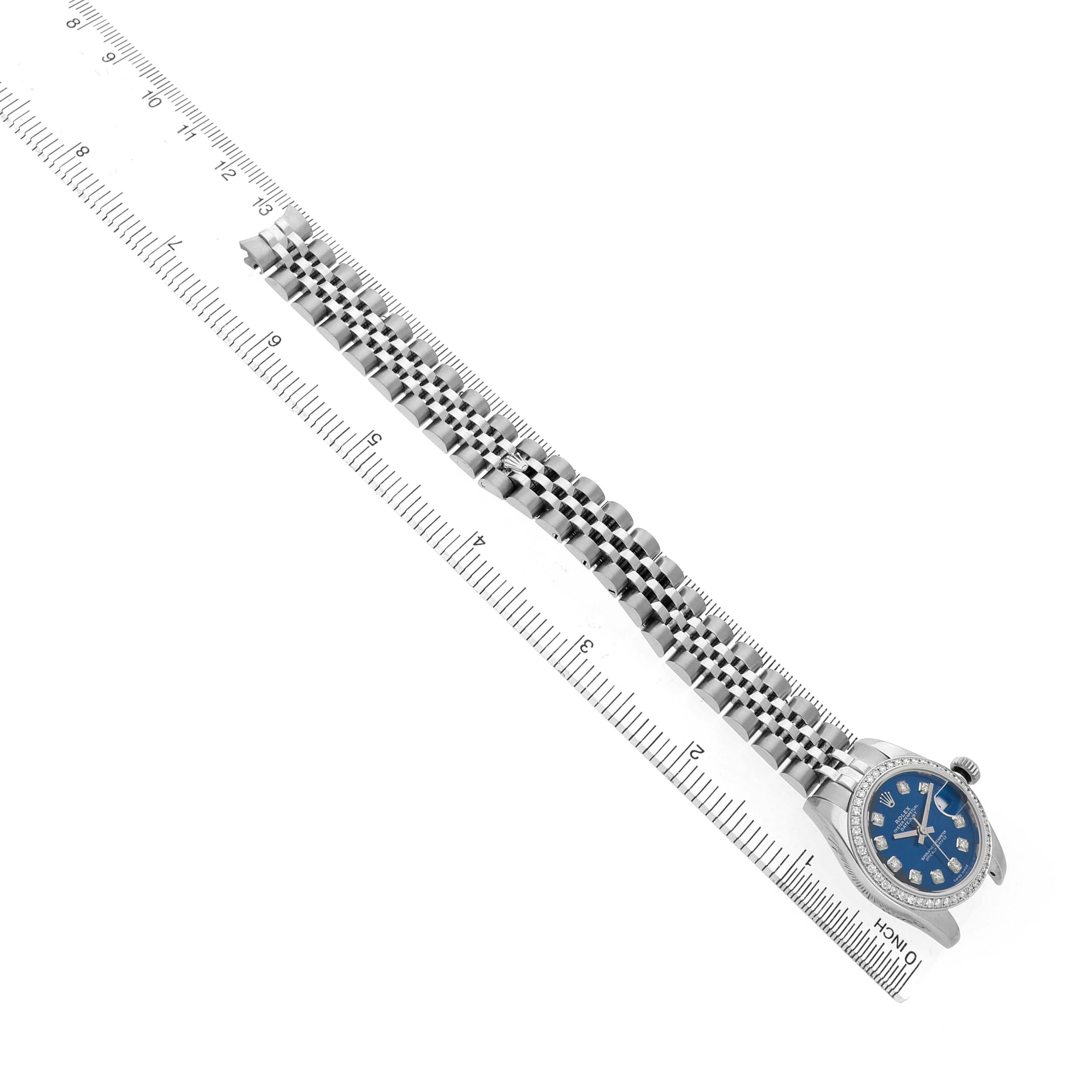 The Rolex Datejust 179384 Women's Stainless Steel Blue Dial watch is displayed diagonally, showing the face, bezel, and full bracelet laid alongside a ruler.