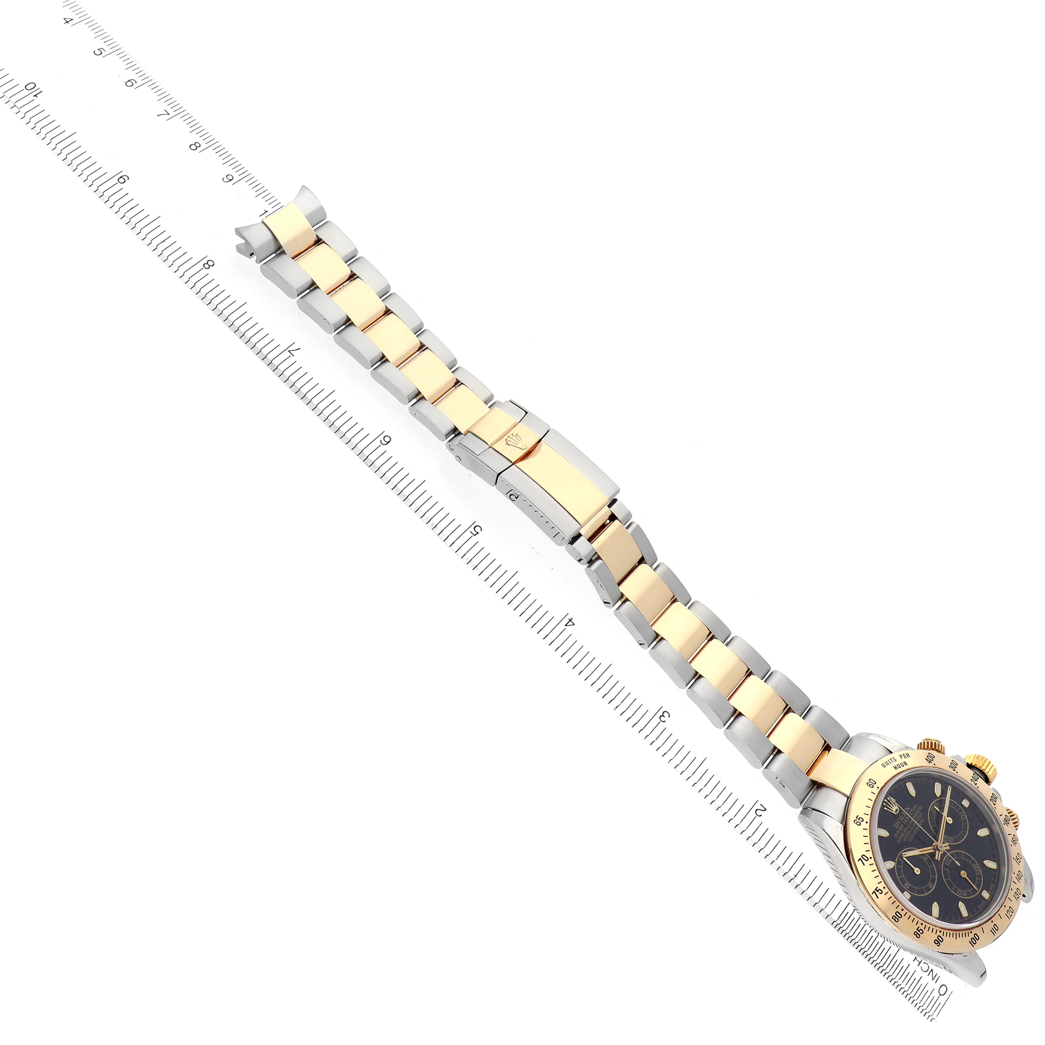 The Rolex Daytona model 116523 men's watch is shown at an angle displaying its gold and silver bracelet, clasp, and black dial, with a ruler for scale.