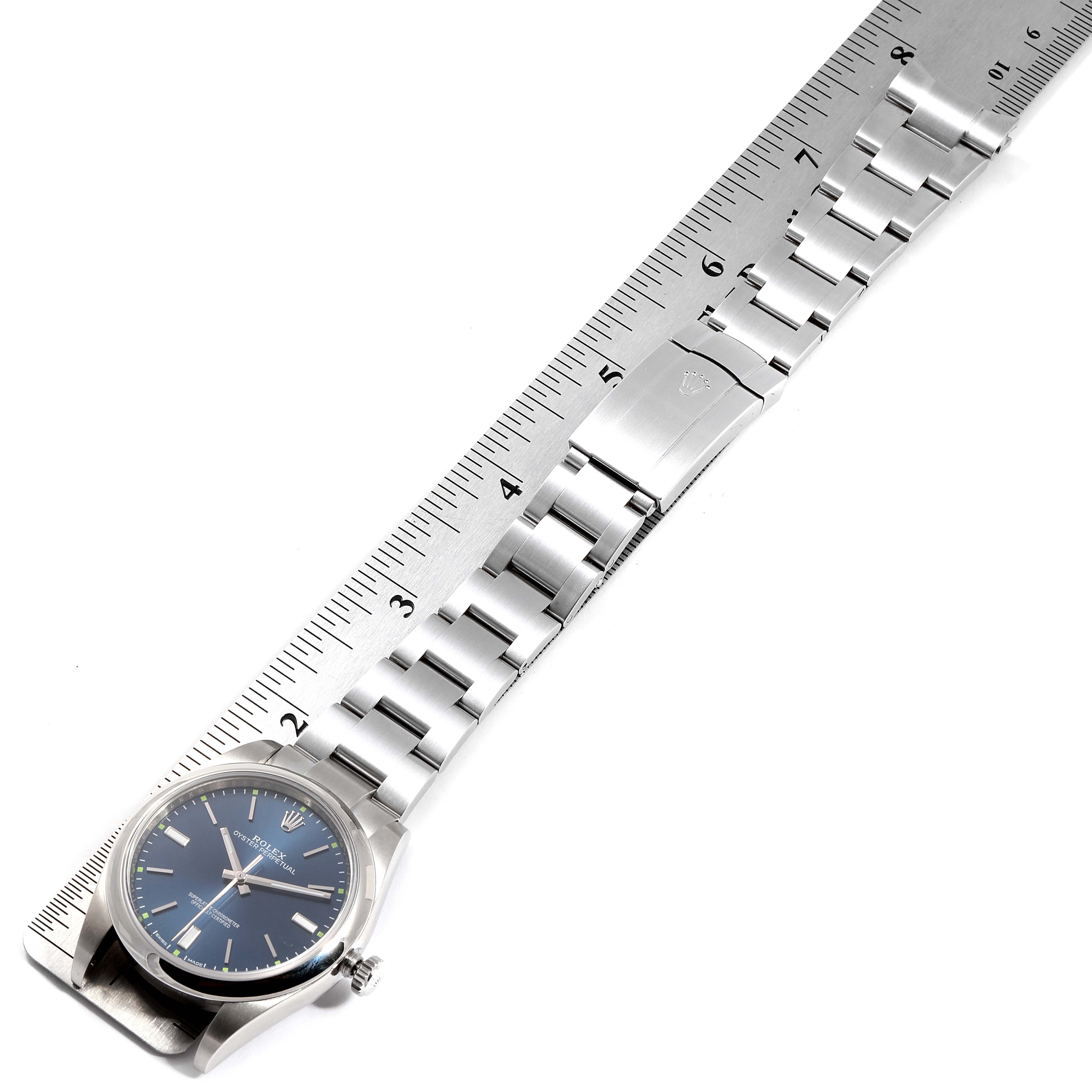This image shows a Rolex Oyster Perpetual 114300 Men's Stainless Steel Blue Dial 114300 Men's Stainless Steel Blue Dial watch laid flat with its band and clasp extended, placed next to a ruler.