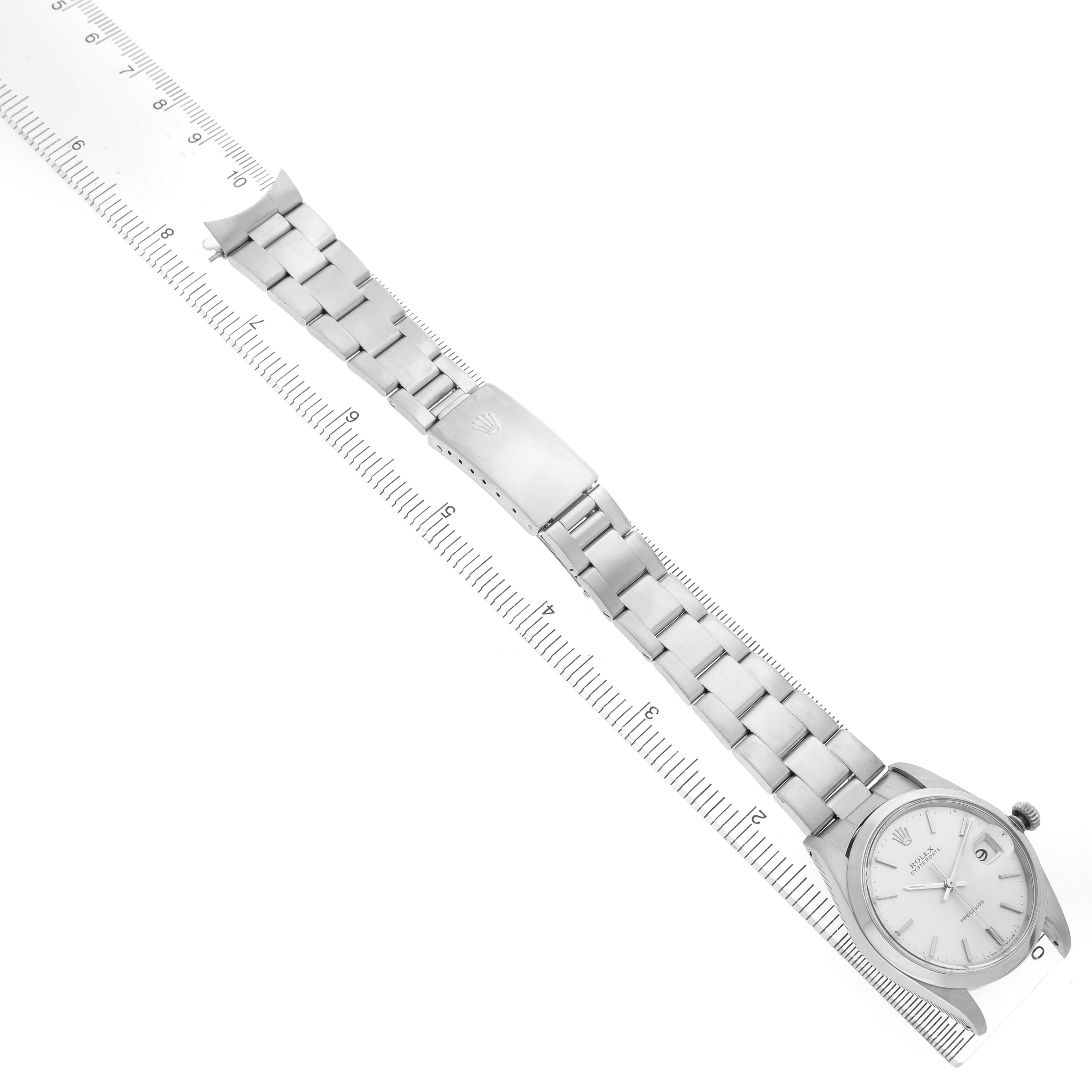 The Rolex Date 6694 Men's Stainless Steel Silver Dial watch is shown from an overhead angle, displaying the face, bracelet, and clasp next to a ruler for scale.