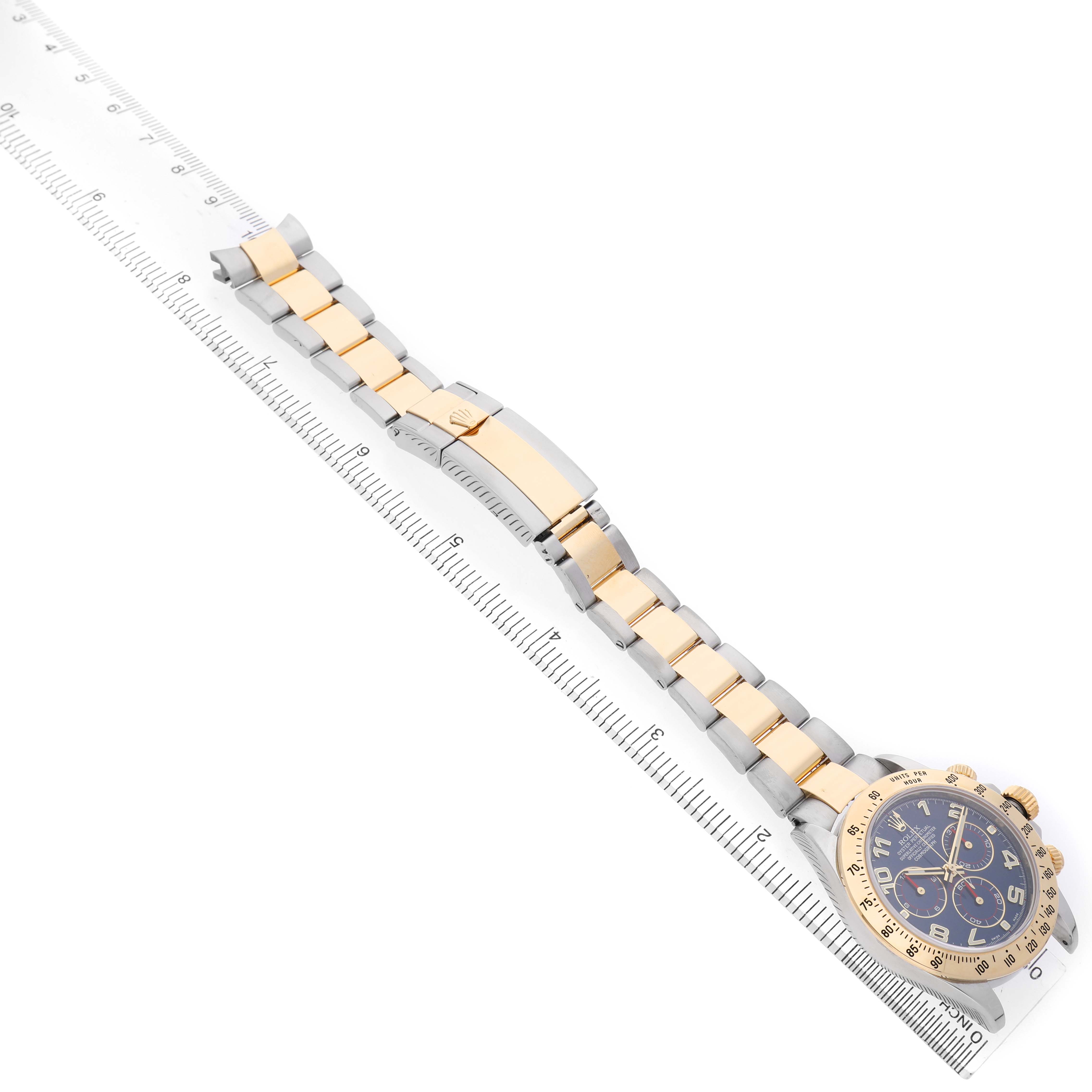 The Rolex Daytona 116523 Men's Steel and Gold (two tone) Blue Dial watch is shown diagonally with its full bracelet, clasp, and face visible alongside a ruler.