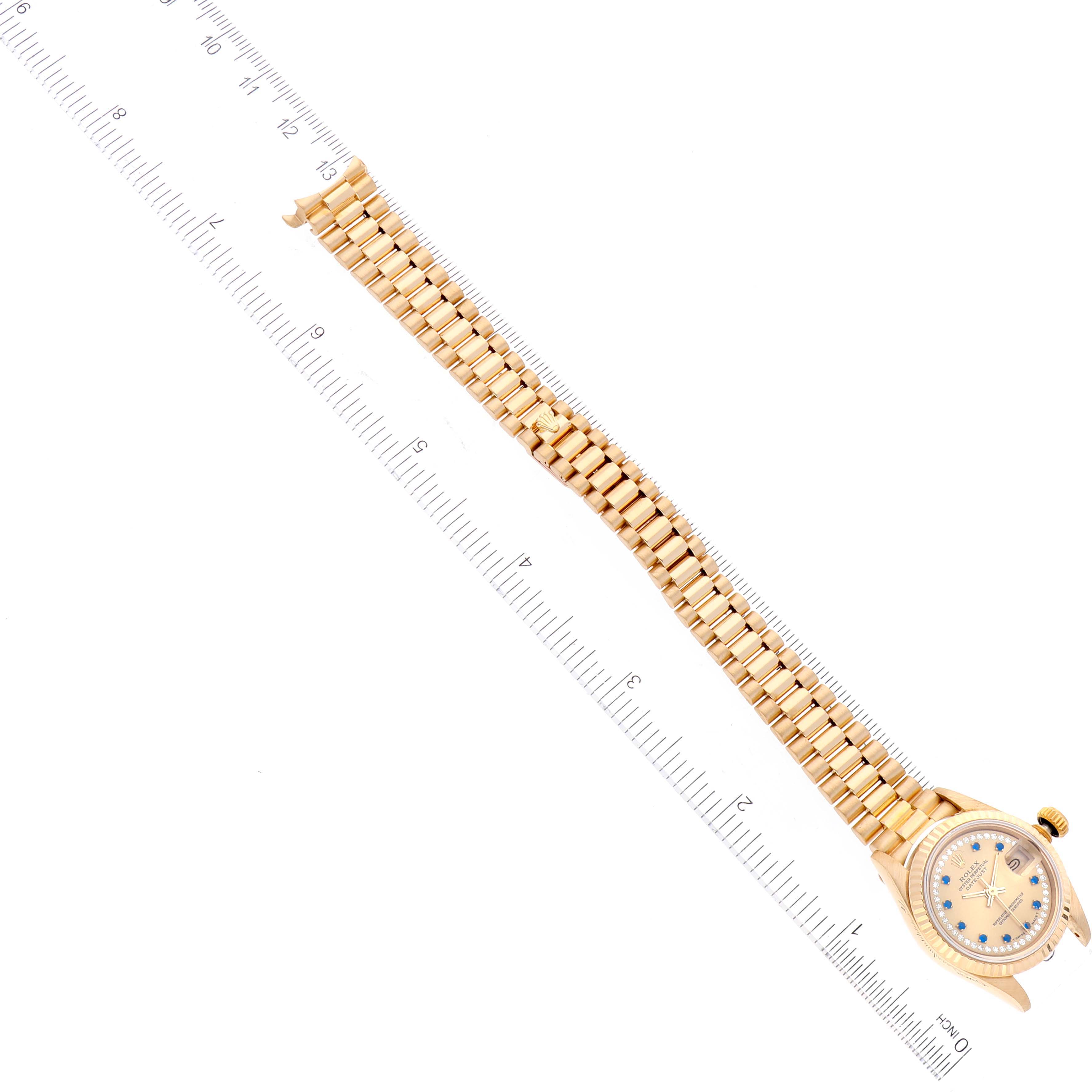 The image shows the Rolex President 69178 Women
s Yellow Gold Champagne Dial 69178 Women
s Yellow Gold Champagne Dial model watch from an elevated angle, displaying its band and face next to a ruler for size reference.