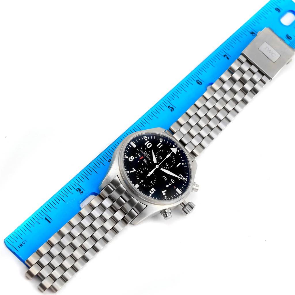 The image shows an IWC Pilot IW377704 Men's Stainless Steel Black Dial IW377704 Men's Stainless Steel Black Dial model watch laying flat, revealing the face, bezel, and bracelet alongside a blue ruler.