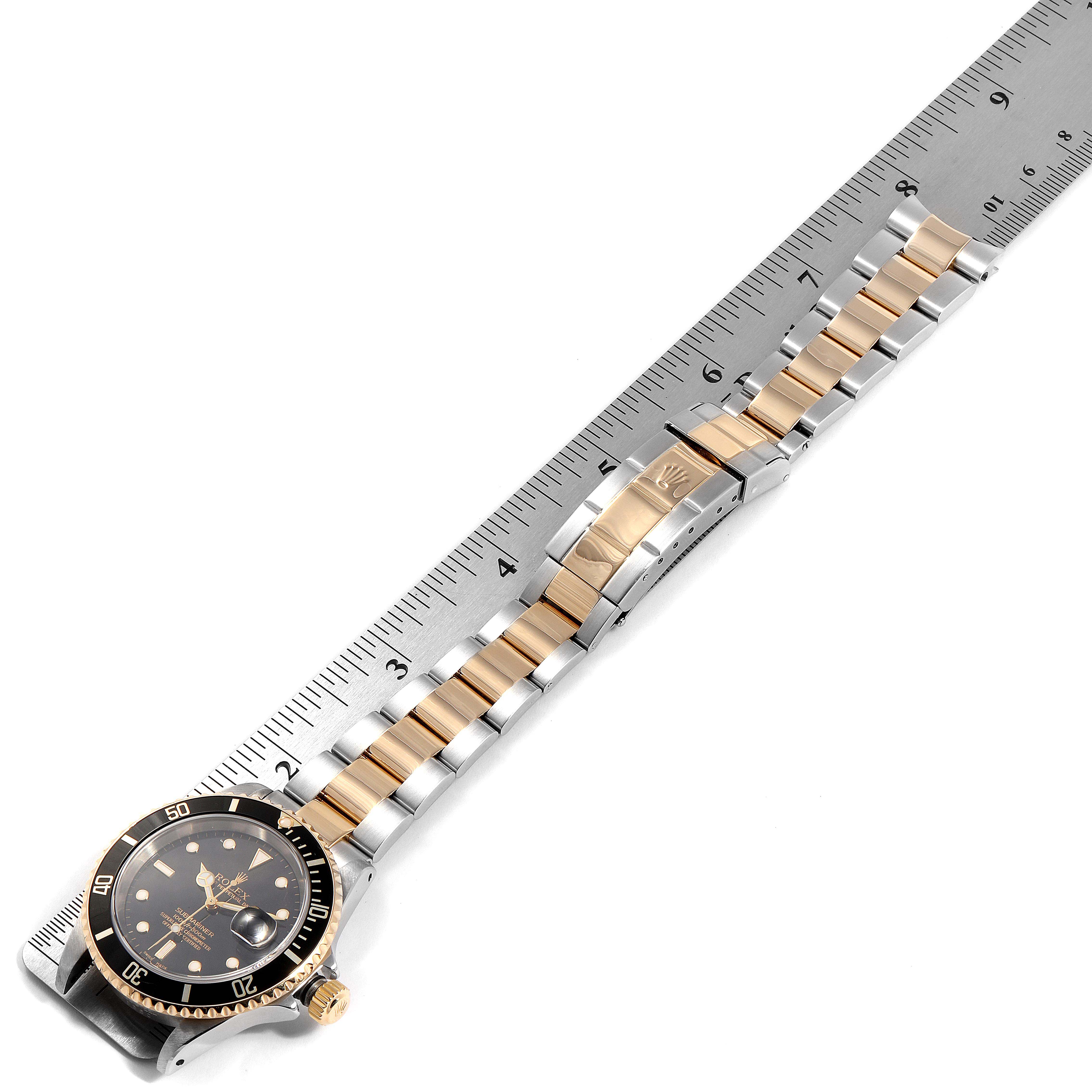 The image shows a Rolex Submariner 16613 Men
s Steel and Gold (two tone) Black Dial 16613 Men
s Steel and Gold (two tone) Black Dial watch placed diagonally on a ruler, displaying its case, bezel, and bracelet.