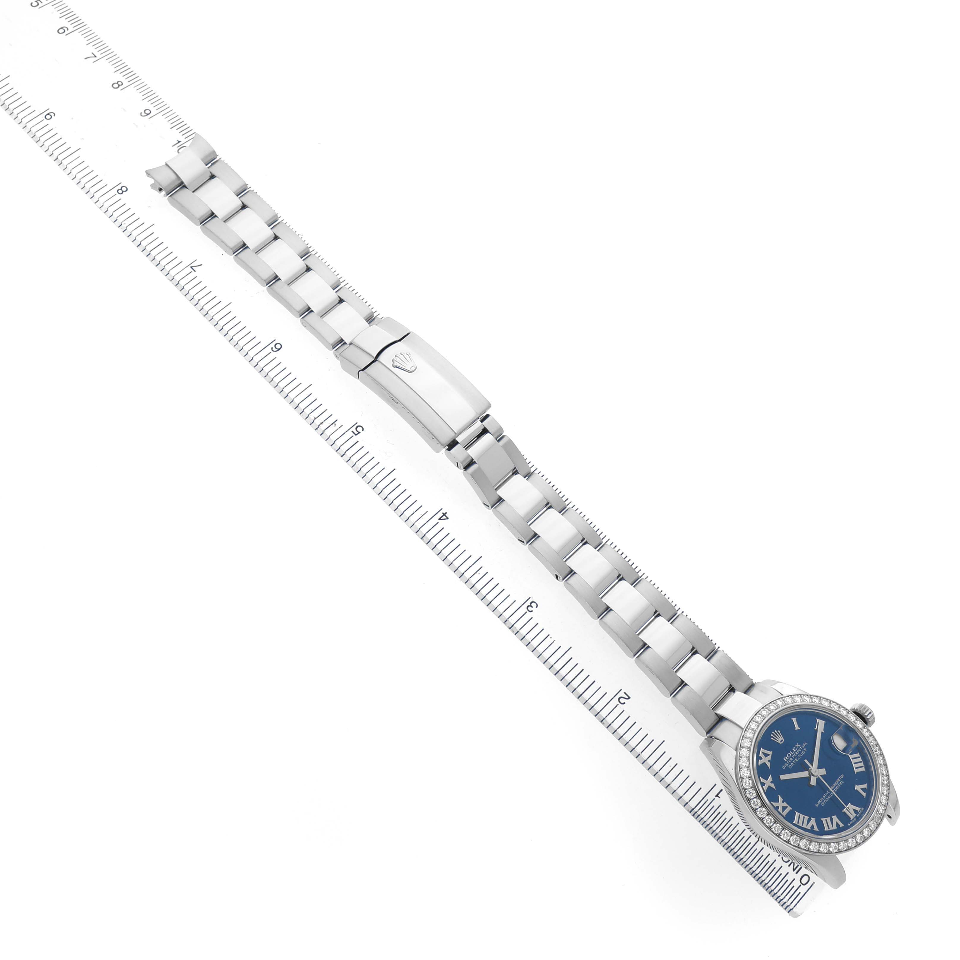 The Rolex Datejust 178384 Women's Stainless Steel Blue Dial watch is shown from a top angle, highlighting its blue dial, Roman numerals, bezel, and bracelet against a ruler.